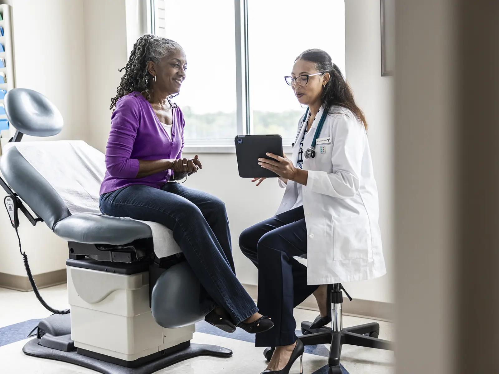 A smiling elderly patient in a purple sweater sits on an exam table, discussing health matters with a doctor using a tablet.