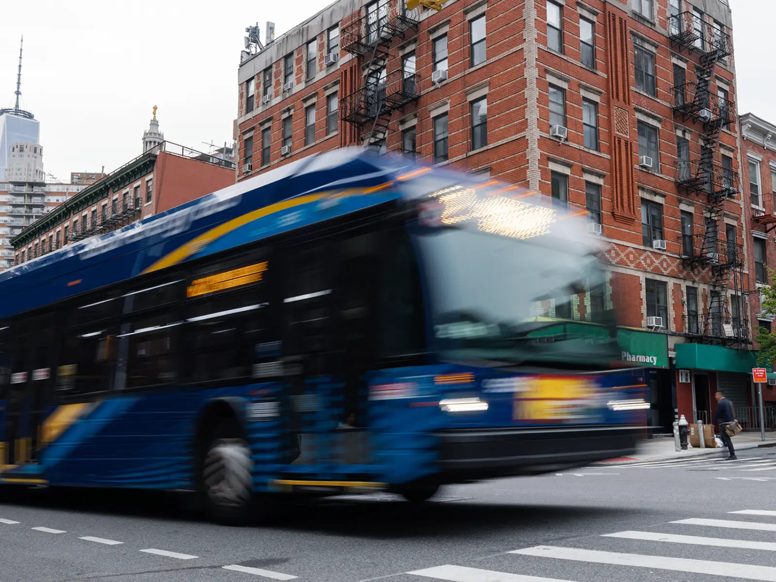 A blue bus speeds through an urban intersection, with red-brick buildings and tall skyscrapers in the background, under a cloudy sky.