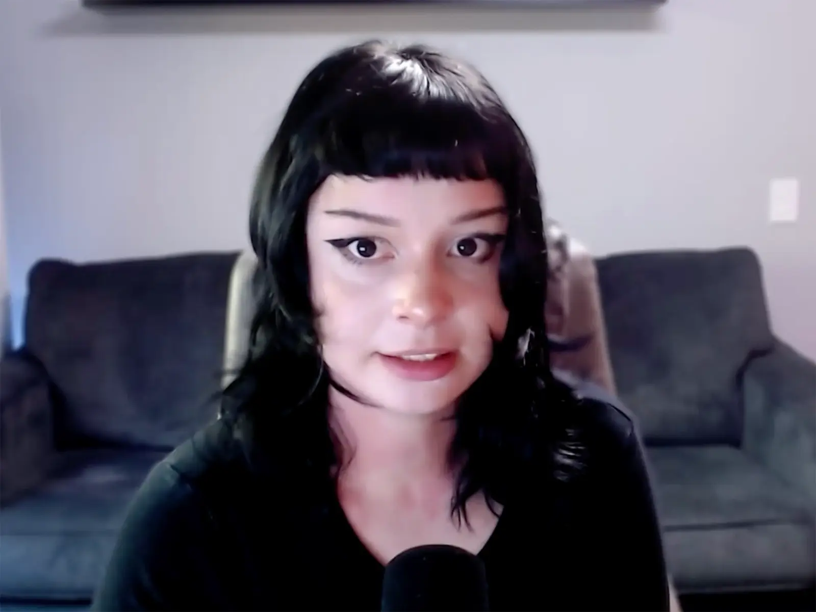 Person with black hair and blunt bangs sits indoors on a couch, facing the camera with a microphone visible in the foreground.