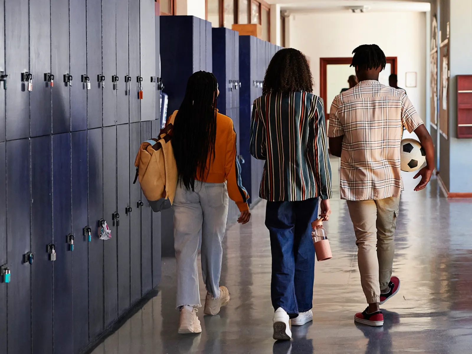 Three students walk away down a school hallway lined with lockers, carrying a backpack, a water bottle, and a soccer ball, seen from behind.
