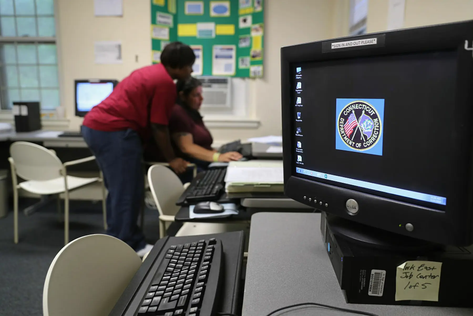 A computer monitor is seen in the foreground as a woman helps another at a computer in the background.