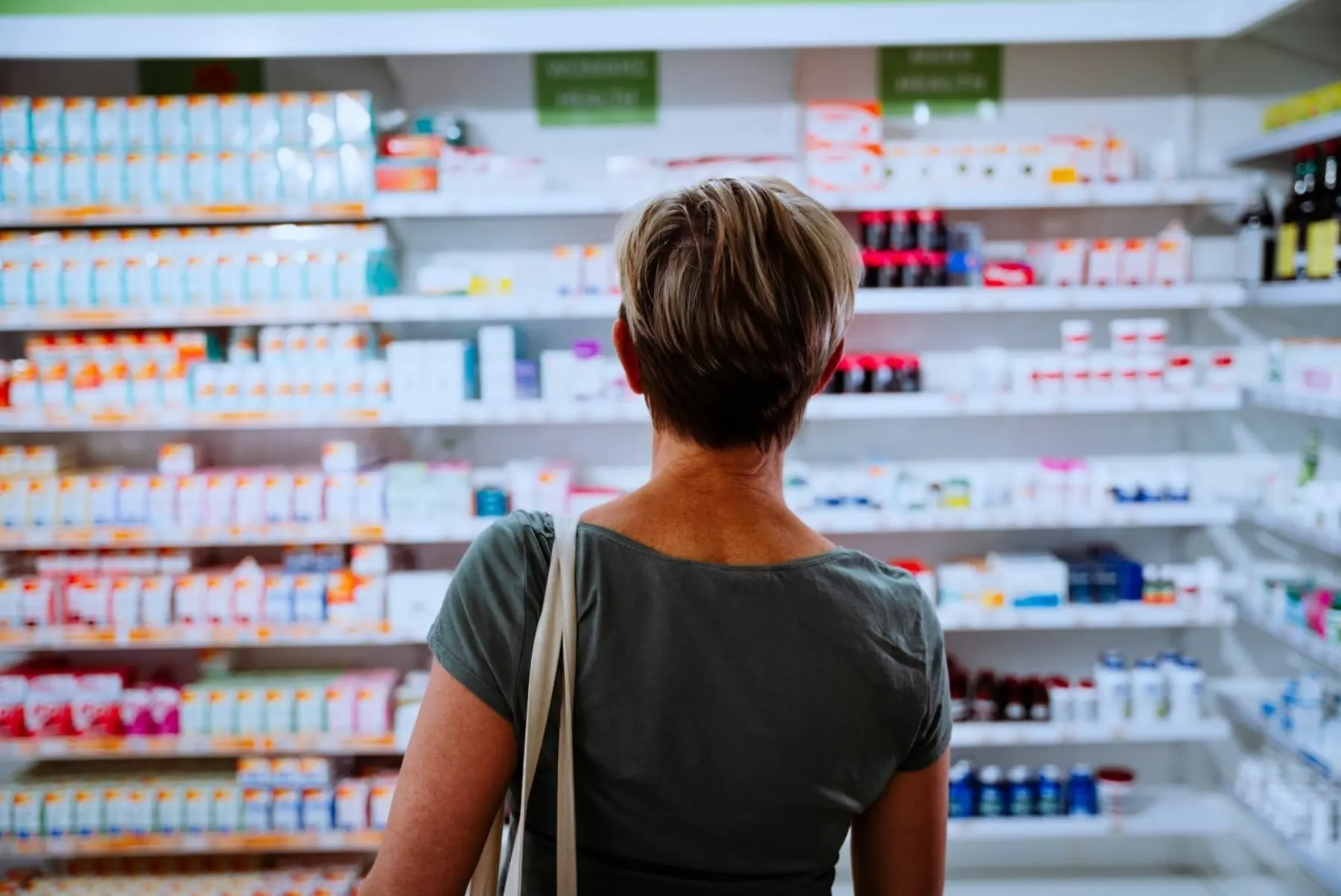 A woman with short hair stands in a pharmacy, gazing at shelves stocked with various medications and health products.