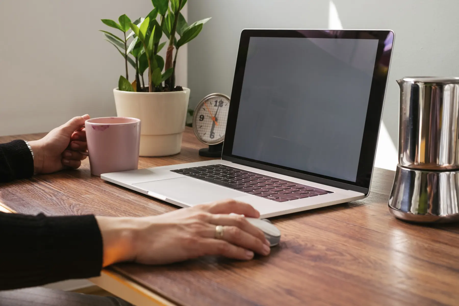 A hand holding a pink mug rests near a laptop on a wooden desk, with a clock and a potted plant in the background.