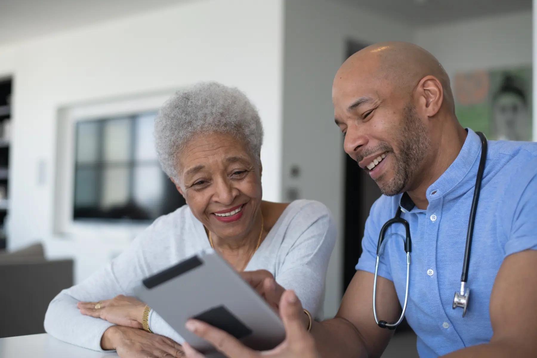 A smiling, older woman and a male doctor share a joyful moment while looking at a tablet in a modern, bright environment.