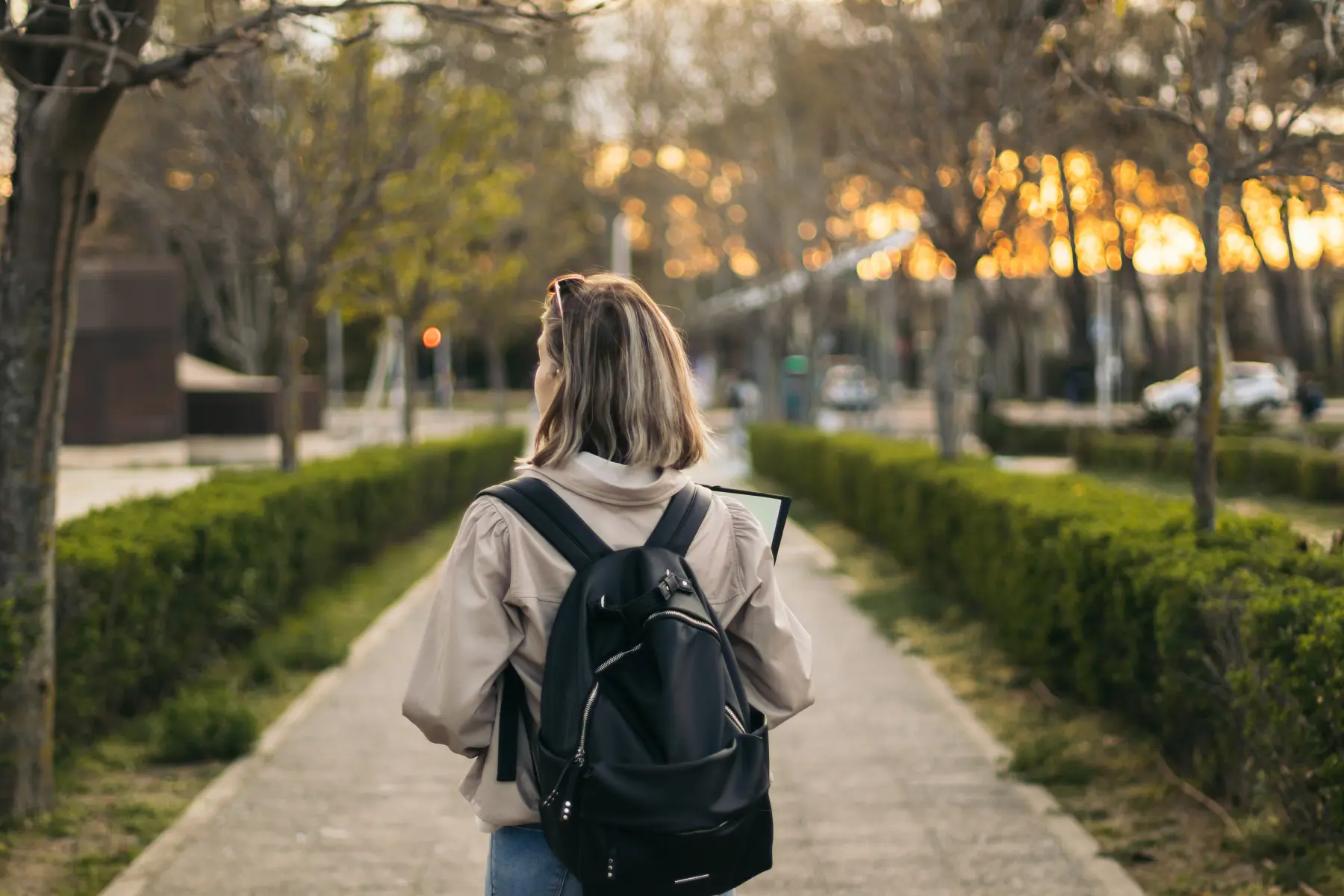 A person walks down a tree-lined path, carrying a black backpack and holding a notebook, with a warm sunset glowing in the background.