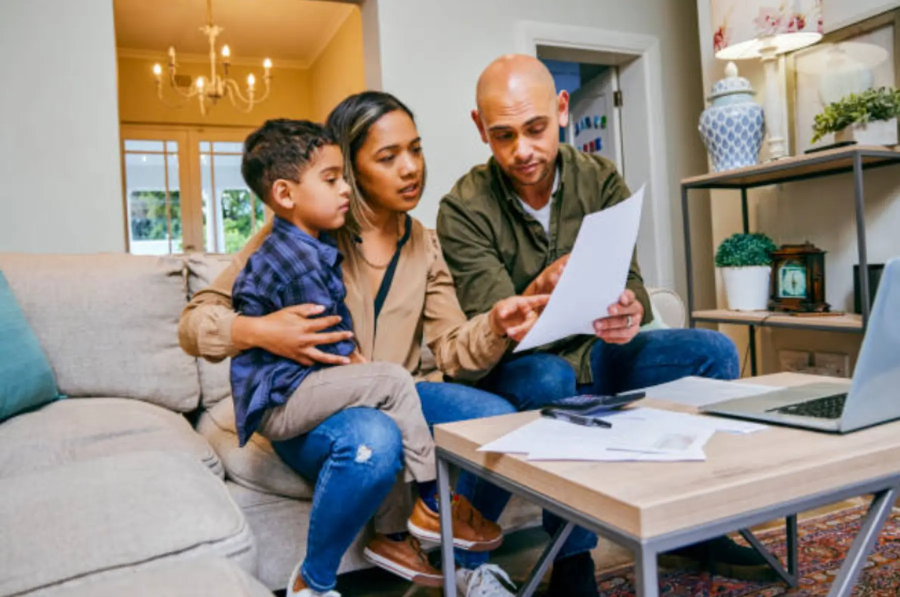 A family of three discusses papers at a coffee table in a cozy living room, with a laptop and documents spread out around them.