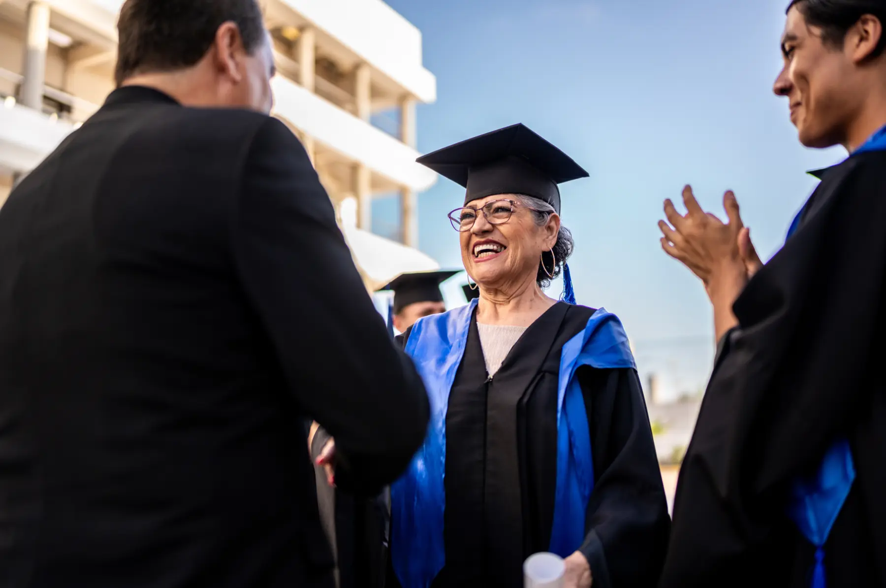 A joyful graduate in a black cap and gown, wearing glasses and hoop earrings, smiles while conversing with a man and others applauding.