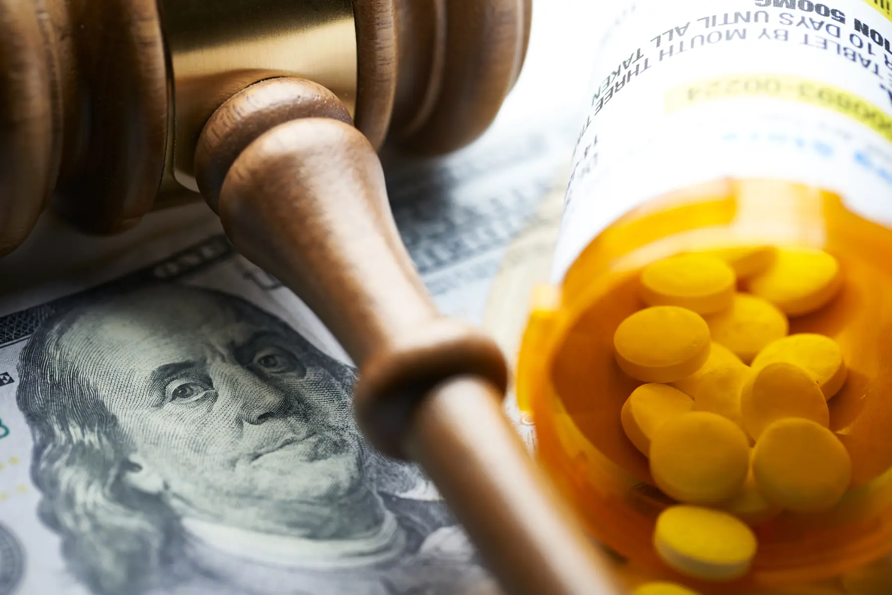 A wooden gavel rests on a hundred-dollar bill alongside an open prescription bottle filled with yellow tablets.