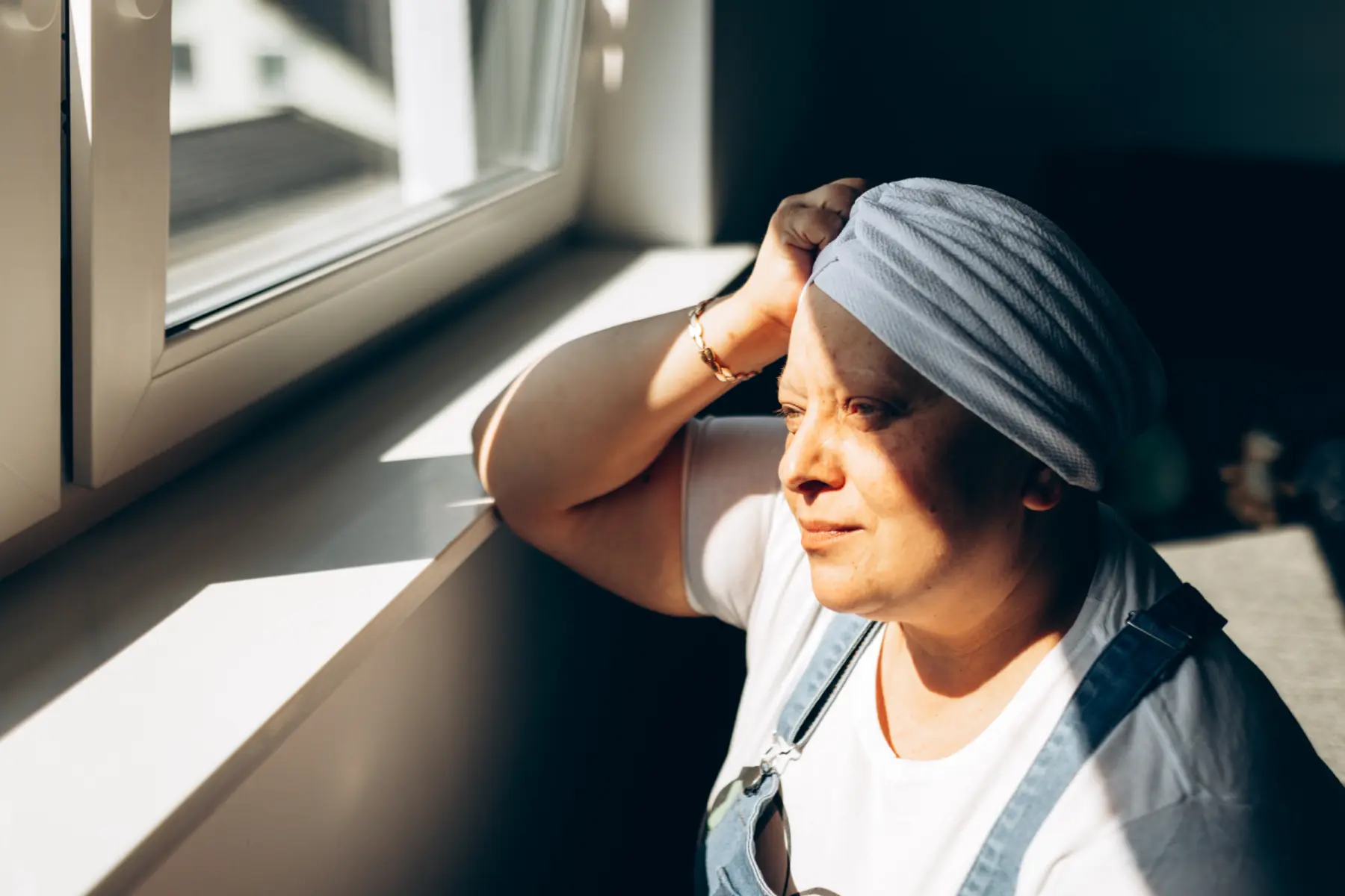 A woman with short hair wrapped in a scarf gazes thoughtfully out of a window, sunlight casting soft shadows around her.