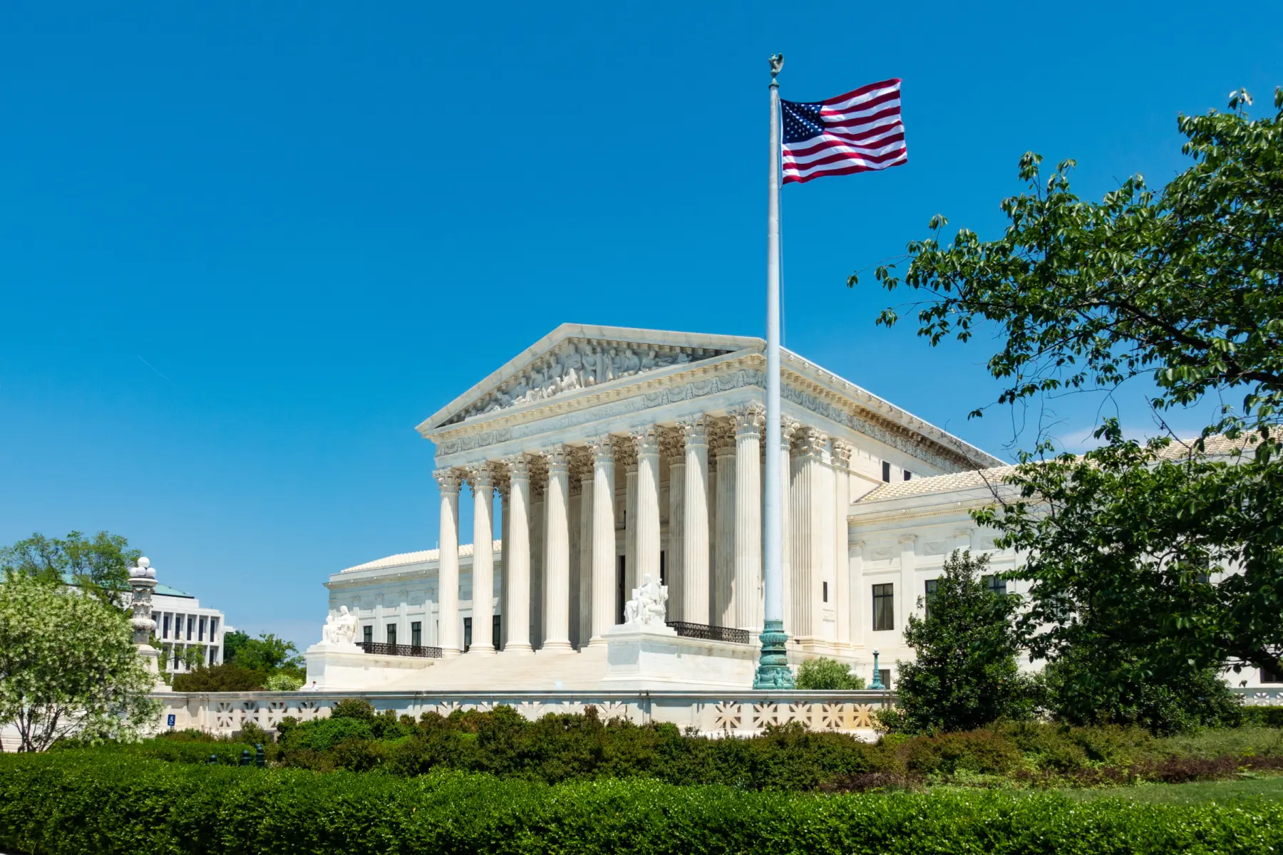 The grand U.S. Supreme Court building features a series of large columns, with a flag waving nearby under a clear blue sky.