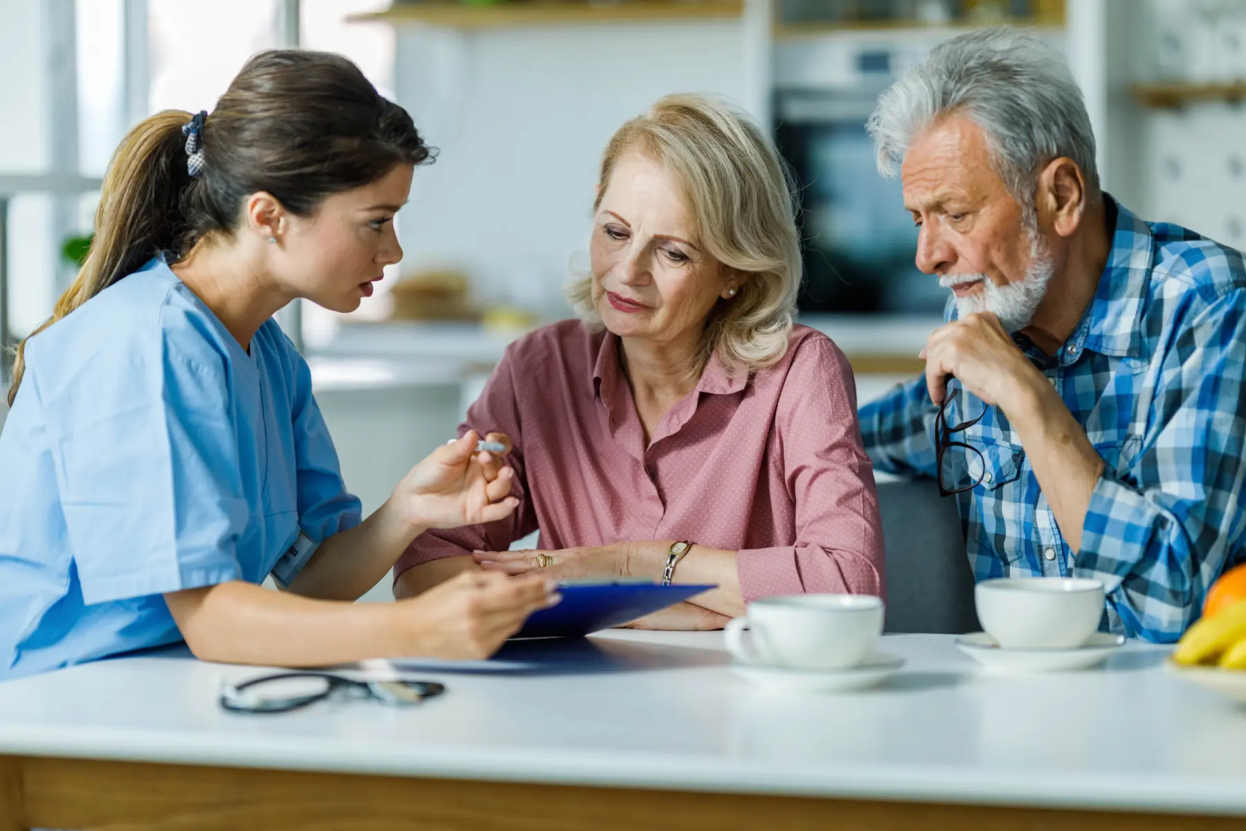A healthcare professional explains information to an elderly couple, seated at a kitchen table with cups and a clipboard visible.