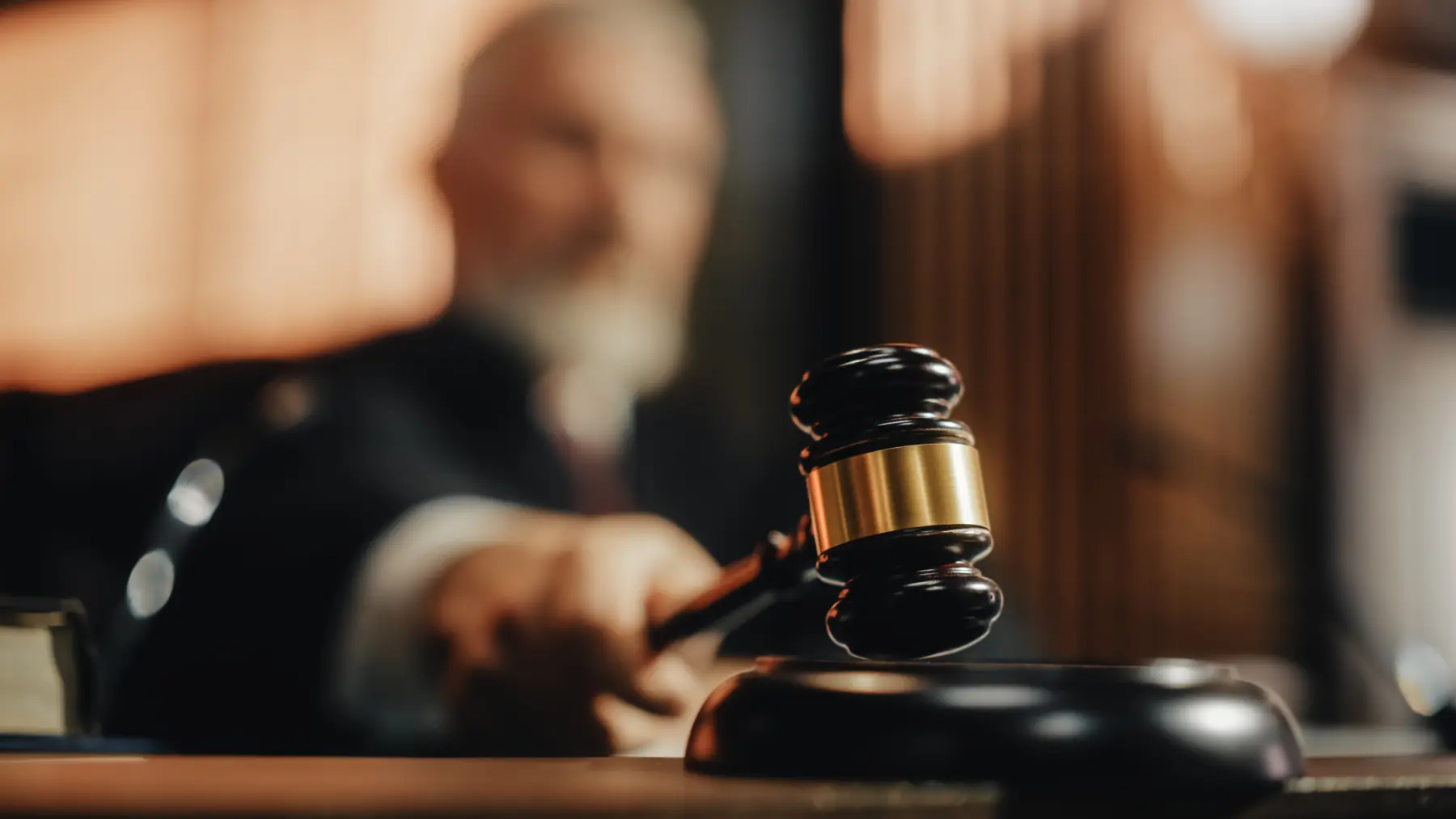 A gavel is poised for a strike on its base, with a blurred judge in the background, evoking a courtroom setting.