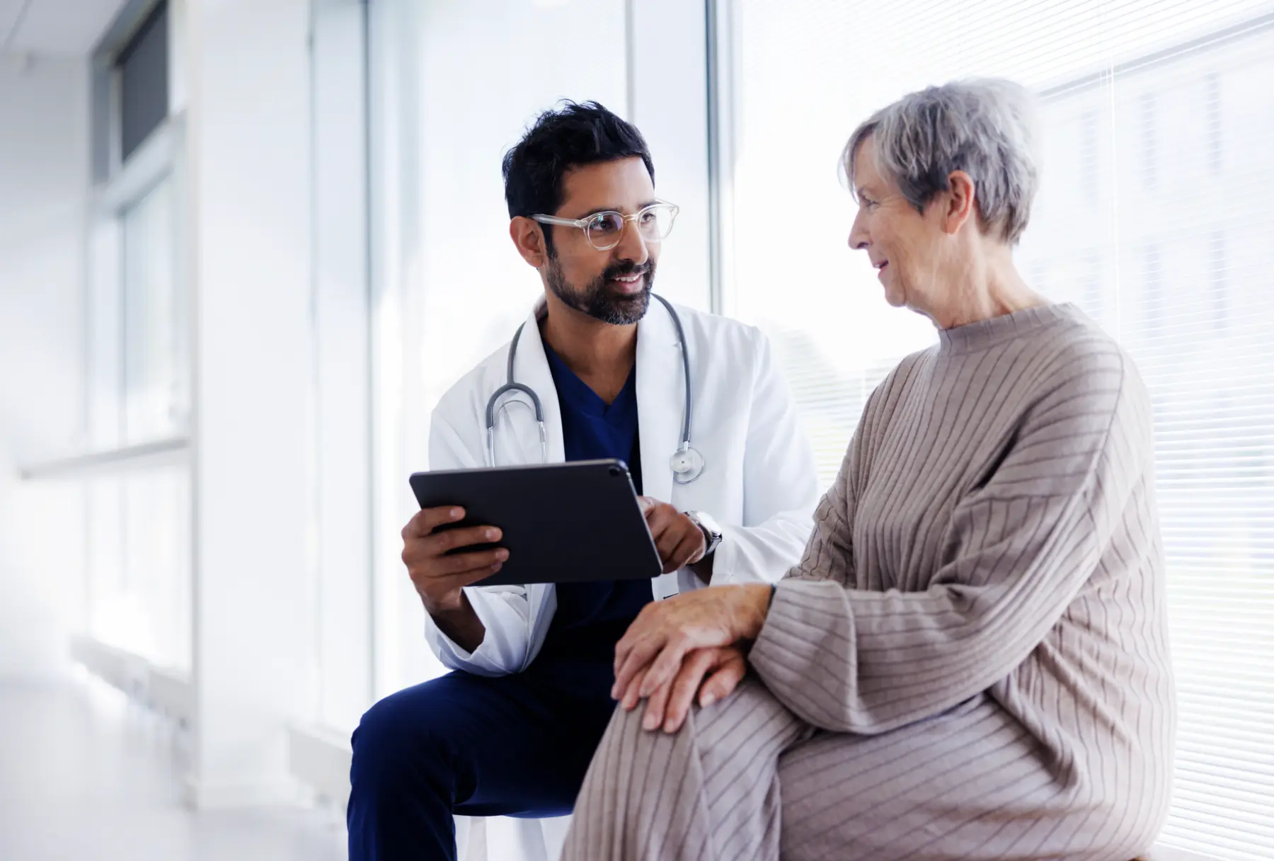 A healthcare professional in a white coat discusses information on a tablet with an older woman, both seated in a well-lit, modern setting.