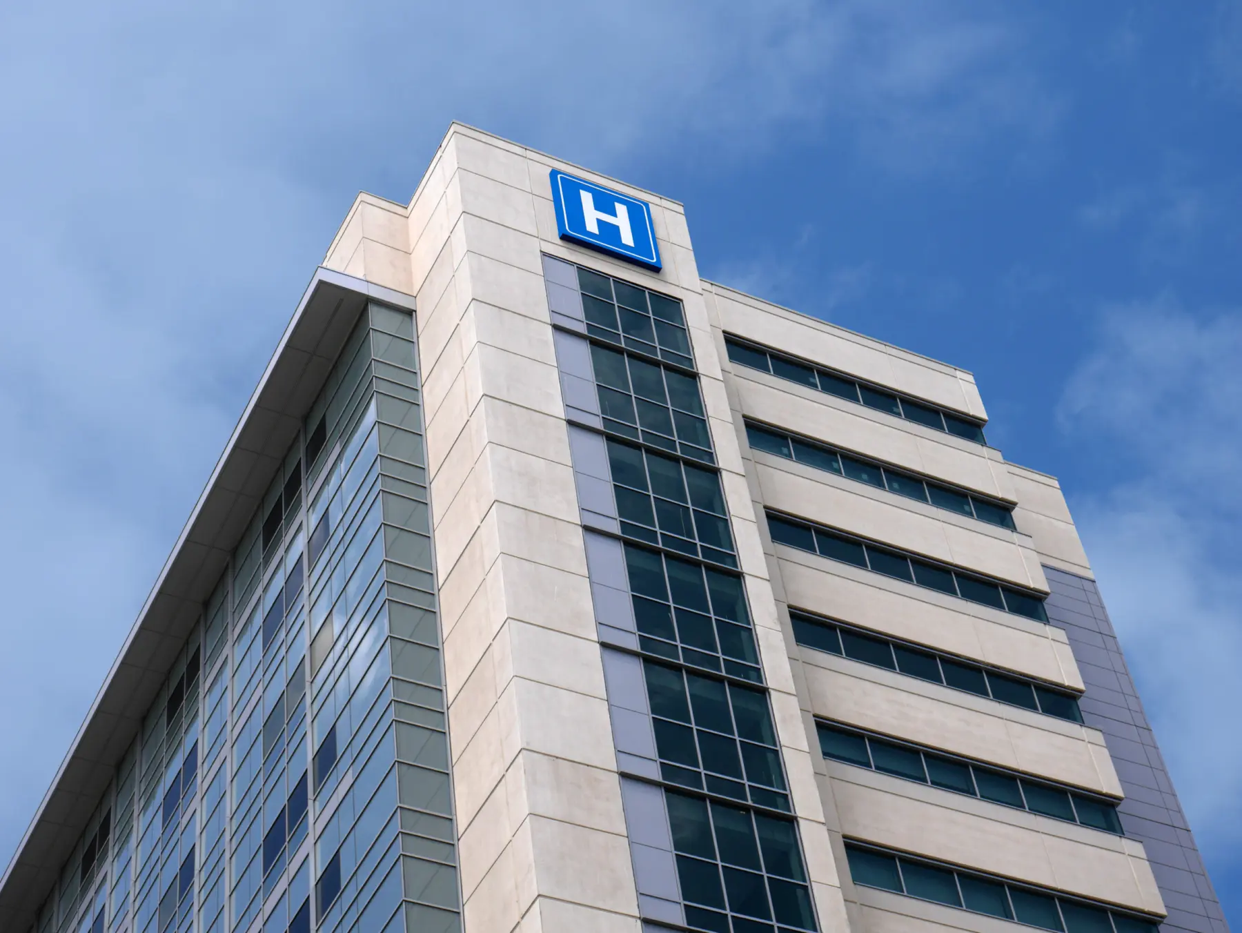 Modern hospital building with large glass windows, a beige façade, and a prominent blue hospital sign against a clear blue sky.