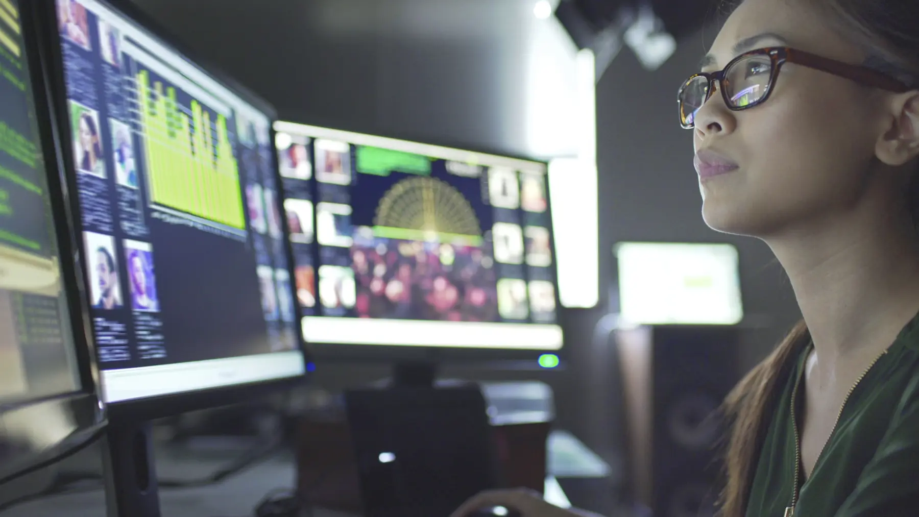 A woman with glasses focuses intently on multiple computer screens displaying data, graphs, and images in a tech environment.