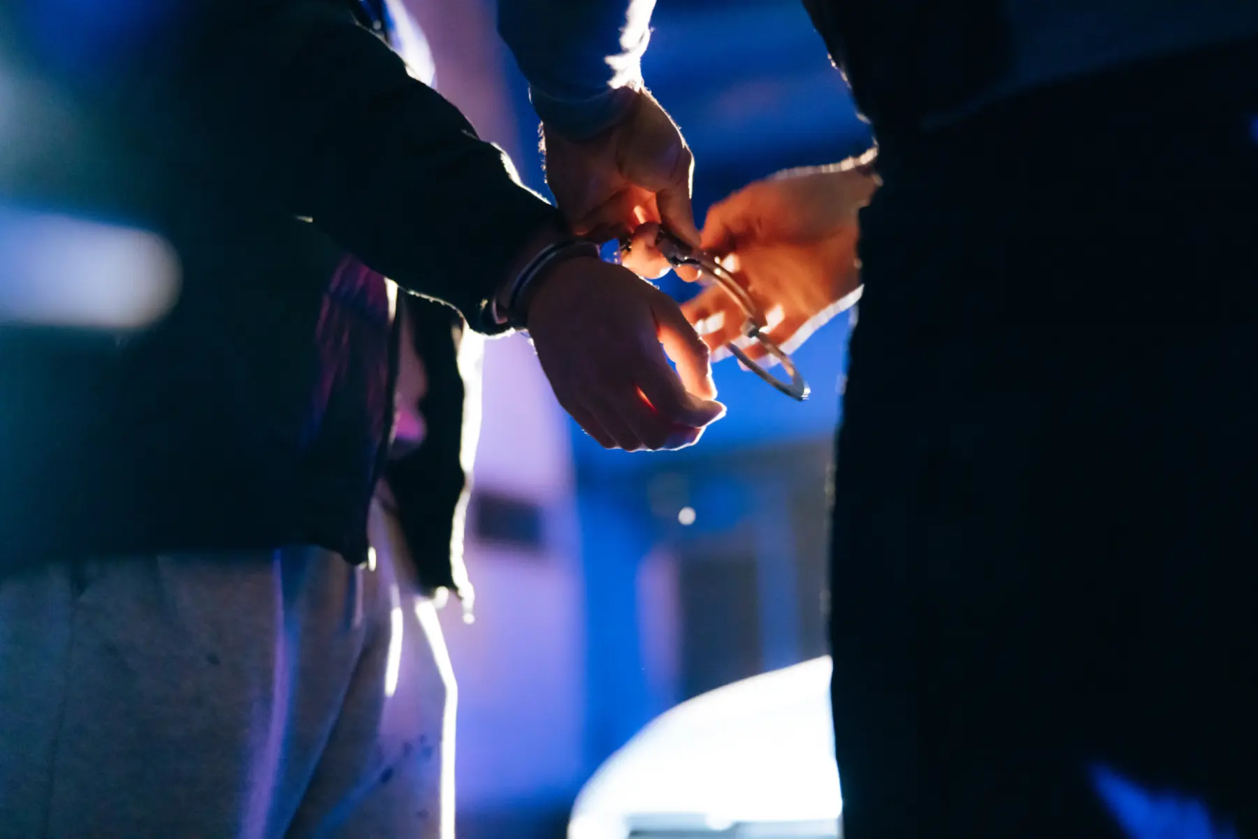Hands are seen fastening handcuffs around a wrist, illuminated by blue and purple lights, suggesting a law enforcement context.
