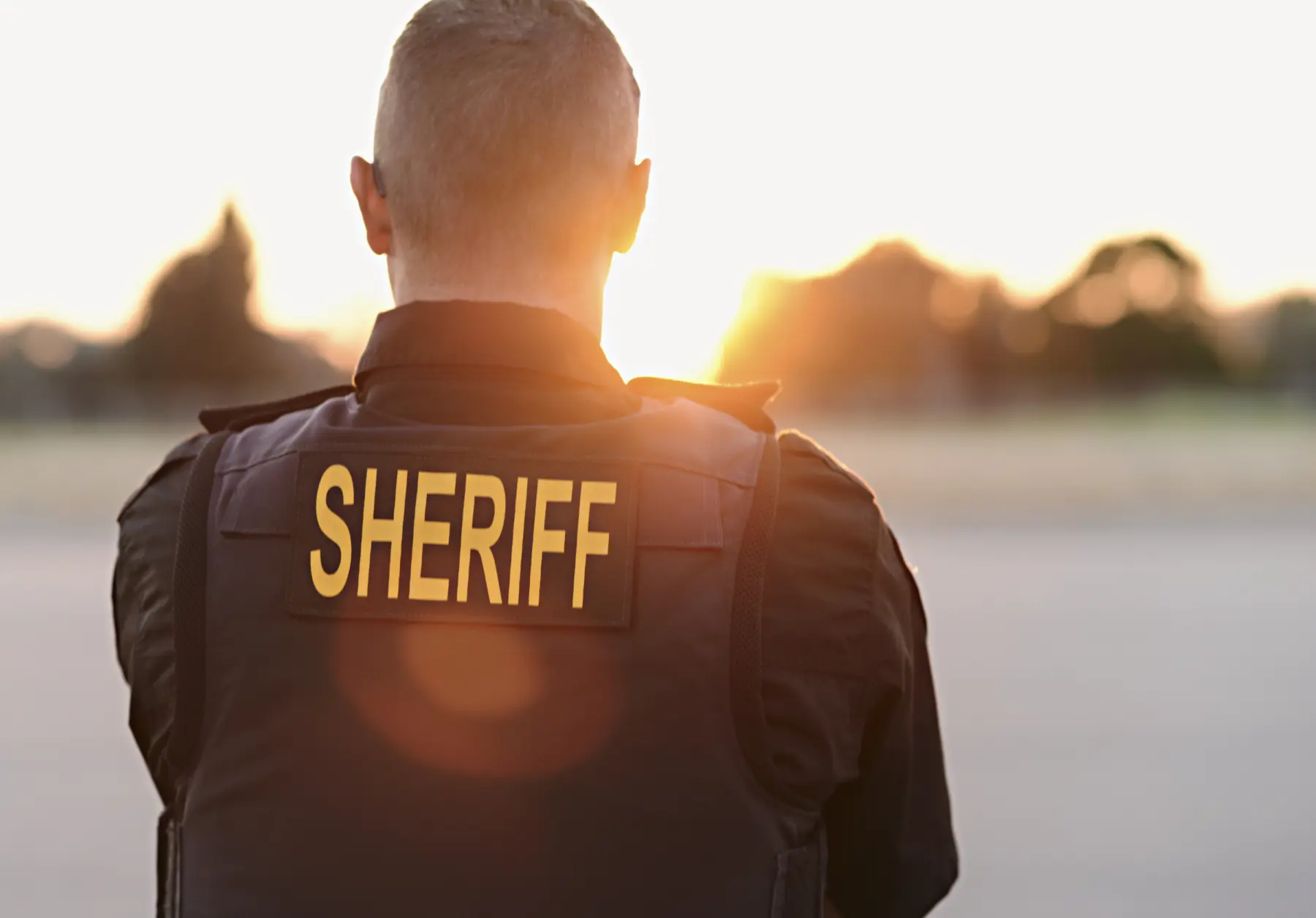 A sheriff stands with their back to the camera, facing a bright sunset, wearing a black vest labeled "SHERIFF" in bold yellow.