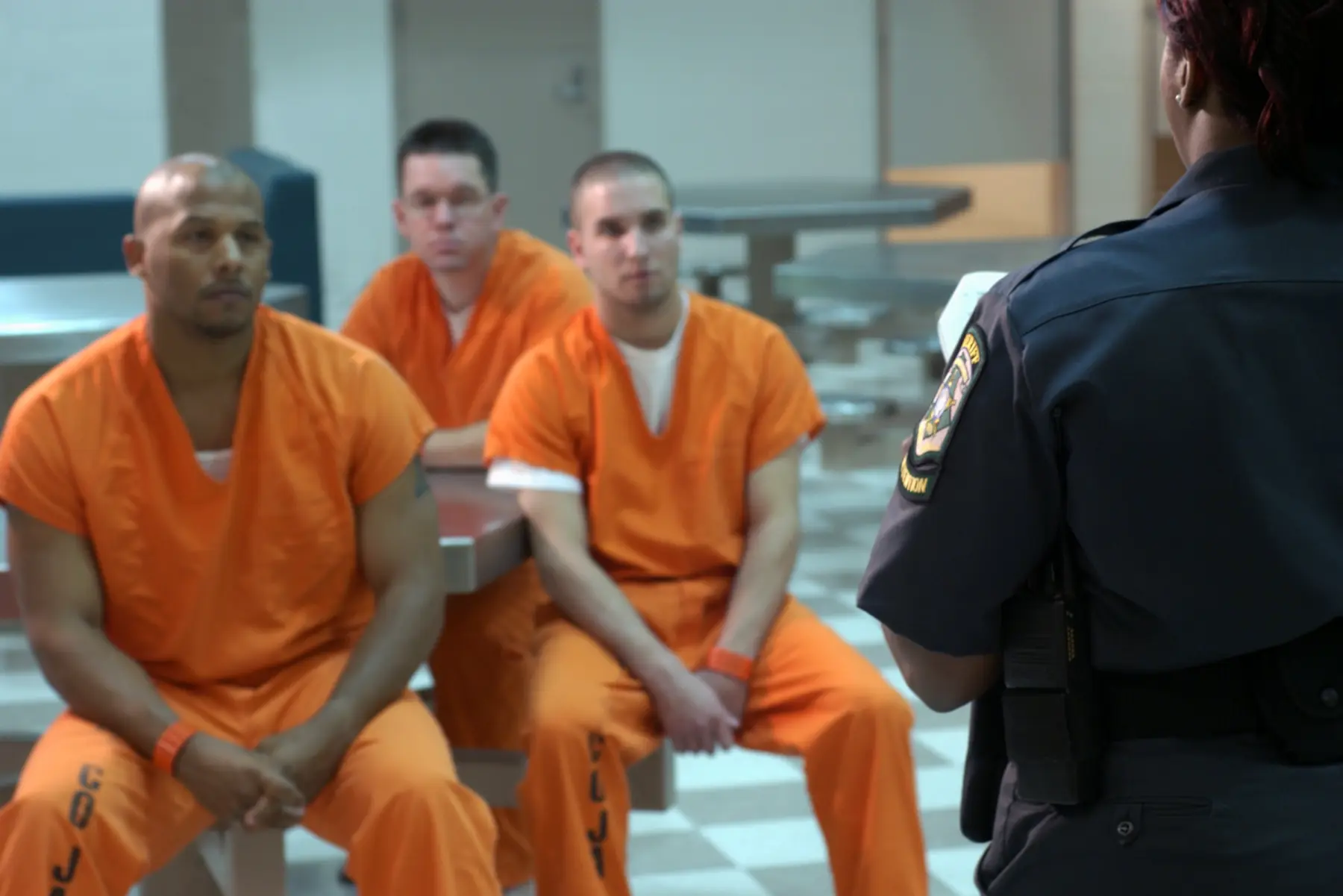 Three male inmates in orange jumpsuits sit on benches, facing a uniformed corrections officer who addresses them in a prison setting.