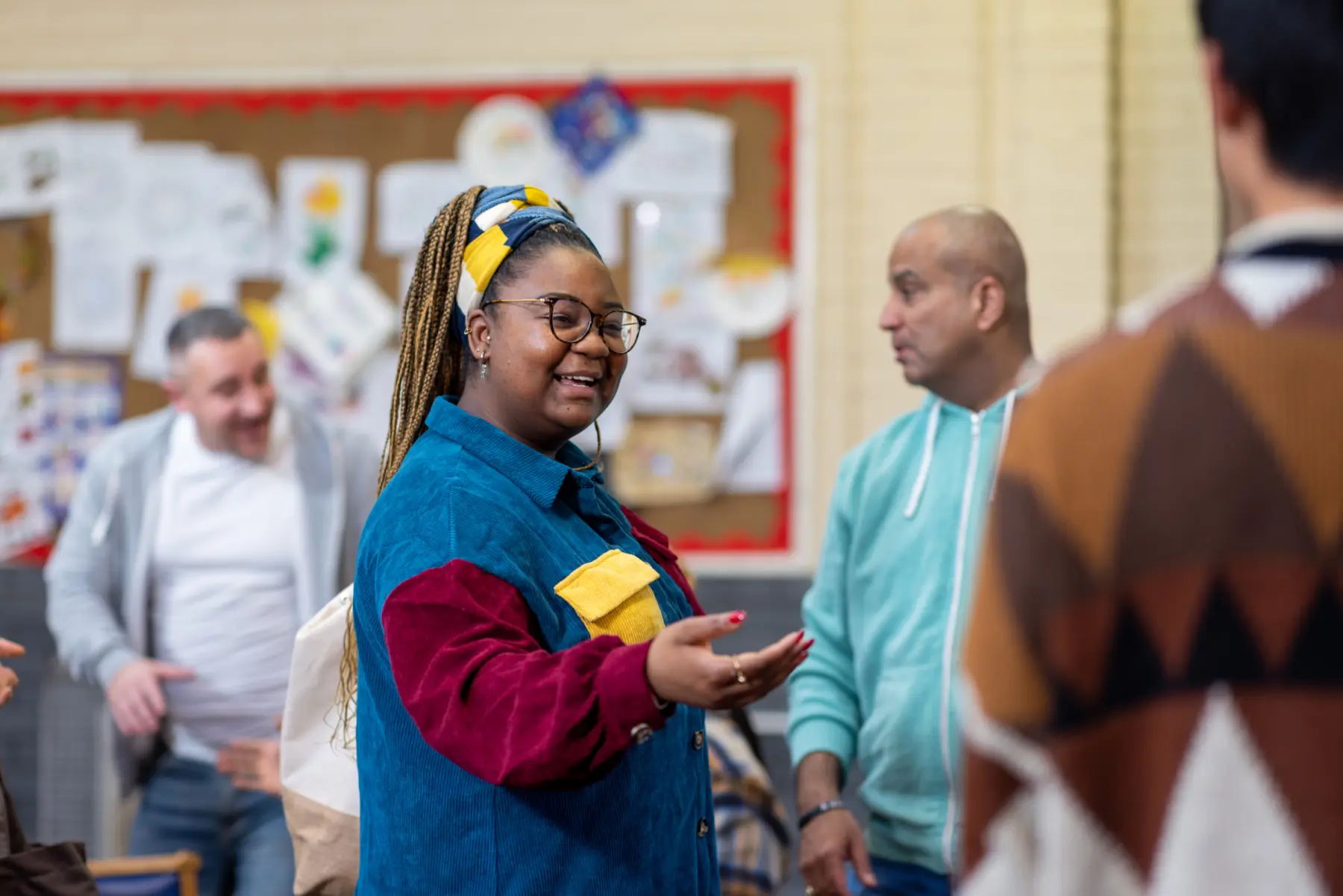 A smiling woman with braided hair, wearing a colorful shirt, engages in conversation with two men in a vibrant community setting.