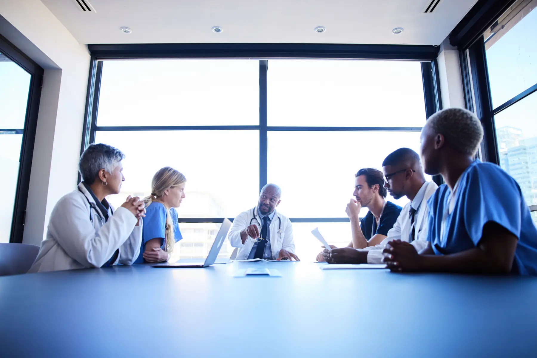 A diverse group of healthcare professionals discusses important topics around a conference table, with large windows in the background.