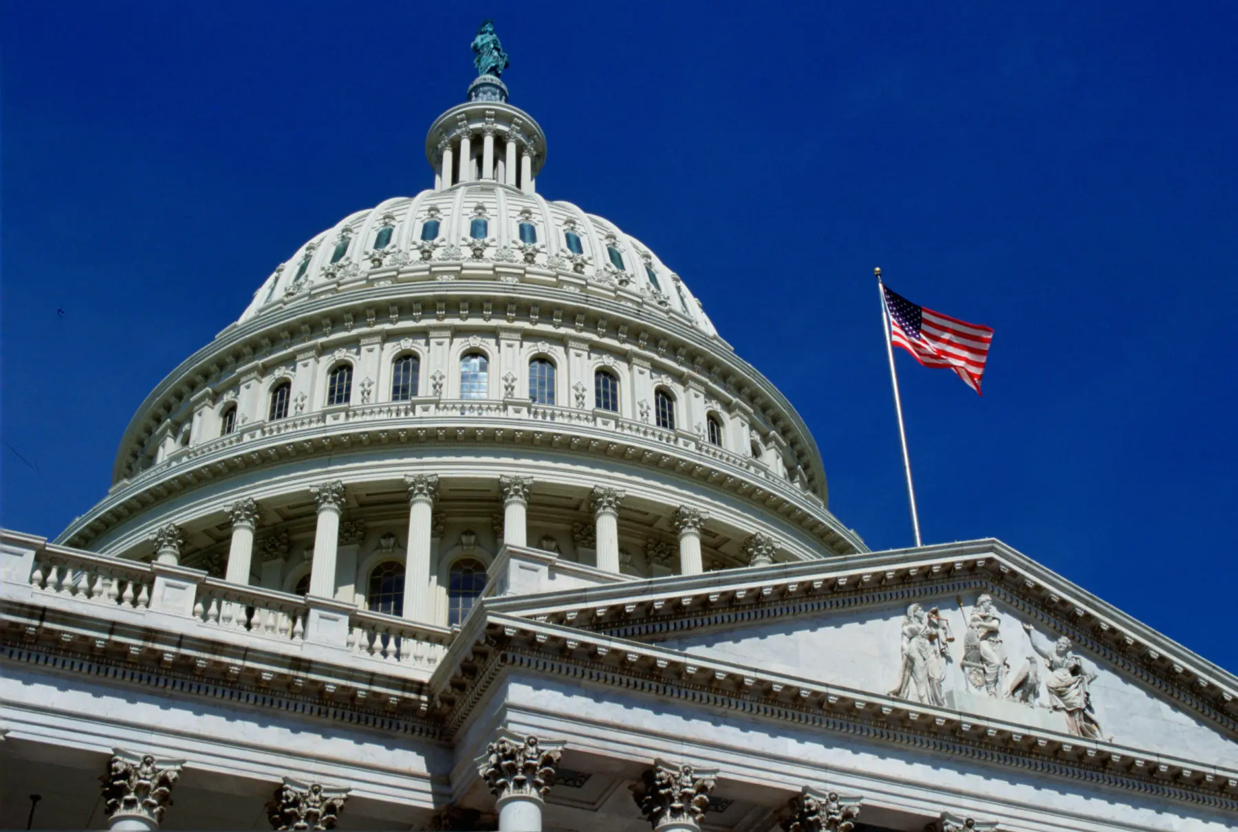 The U.S. Capitol dome rises against a clear blue sky, topped by a statue, with an American flag waving on a columned building.