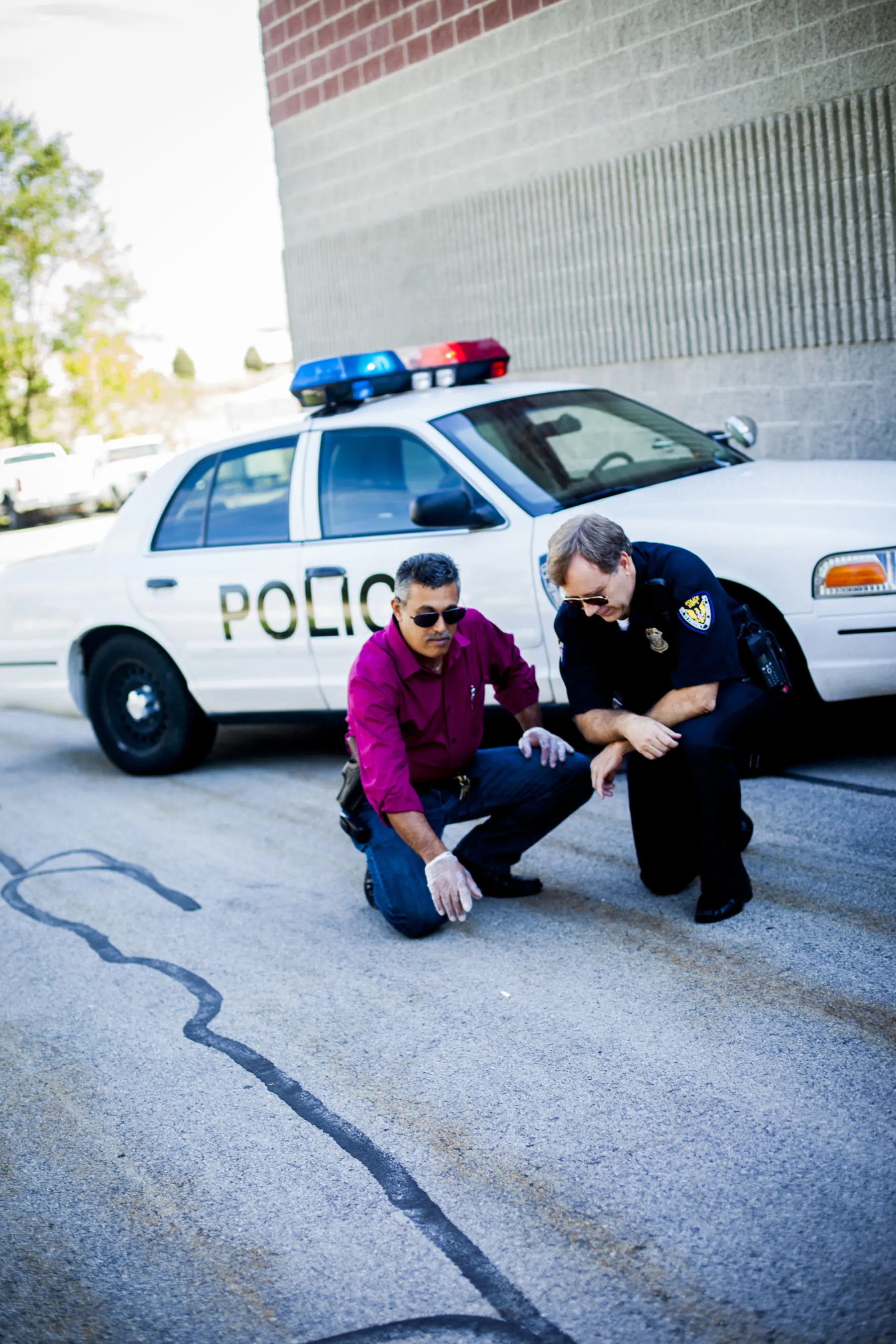 Two men, one in a red shirt and the other in a police uniform, crouch by a marked police car, examining a line on the pavement.