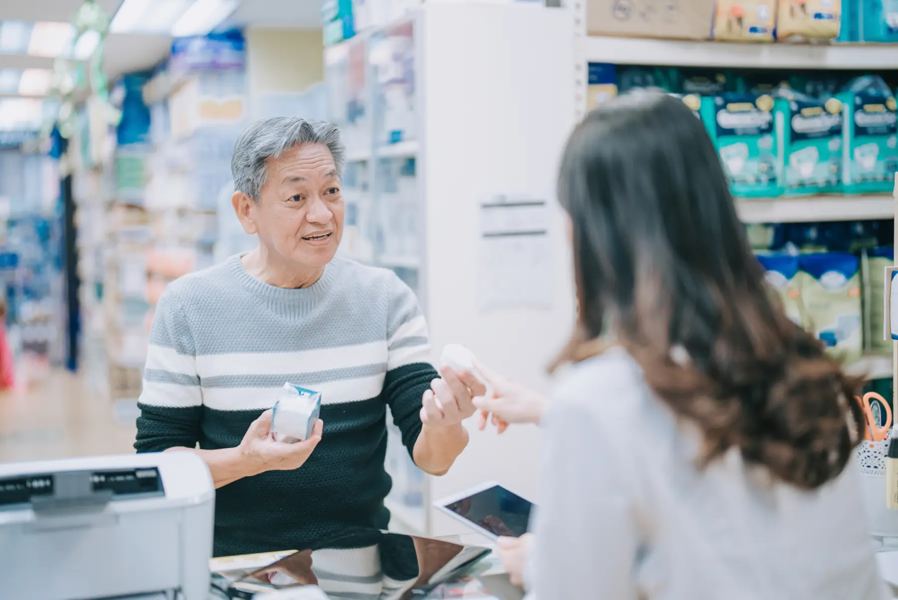 An older man with gray hair smiles while holding a small product, engaging with a woman who is reaching out to receive it.
