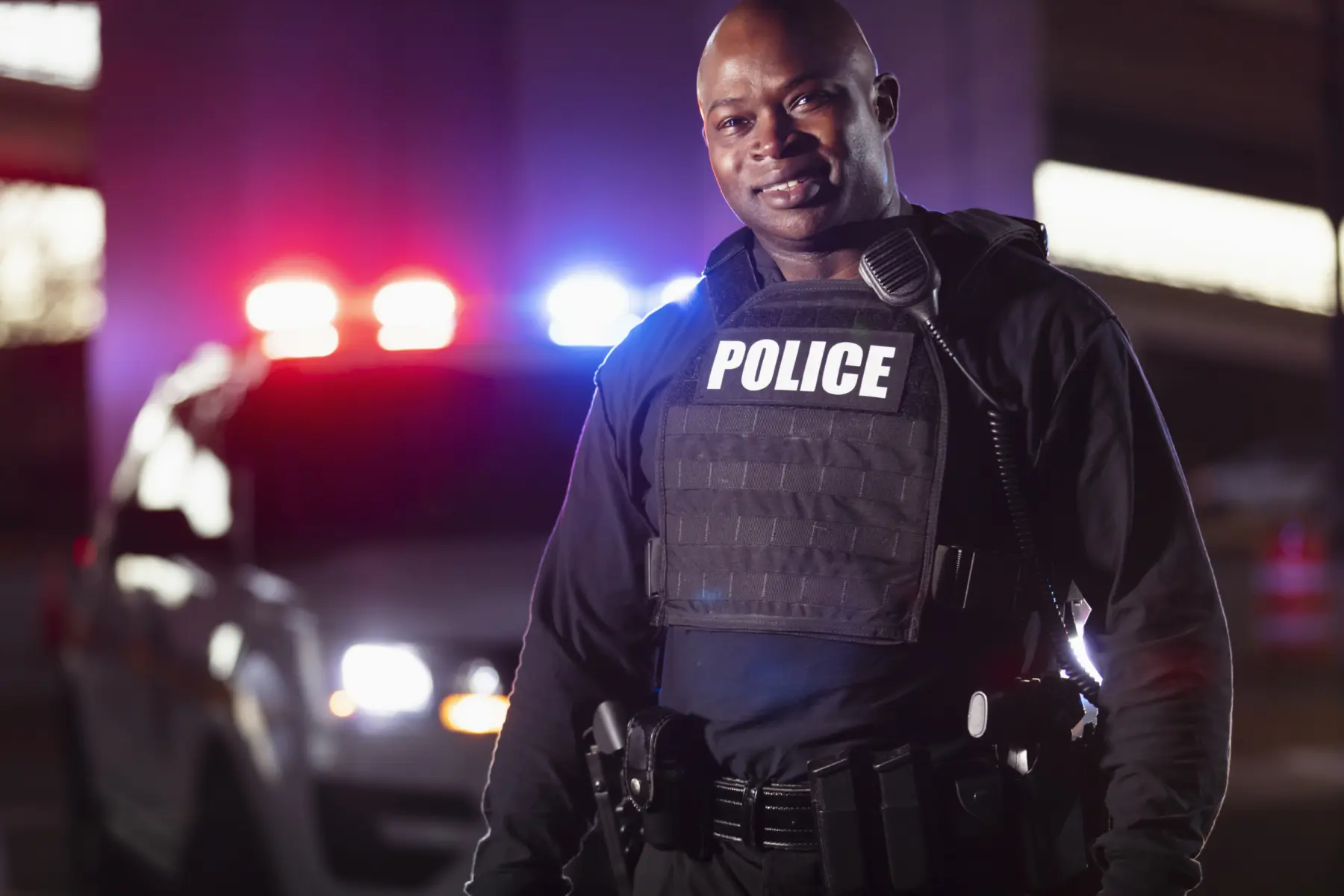 A police officer in a black tactical vest smiles confidently, with a patrol car and flashing red and blue lights in the background.