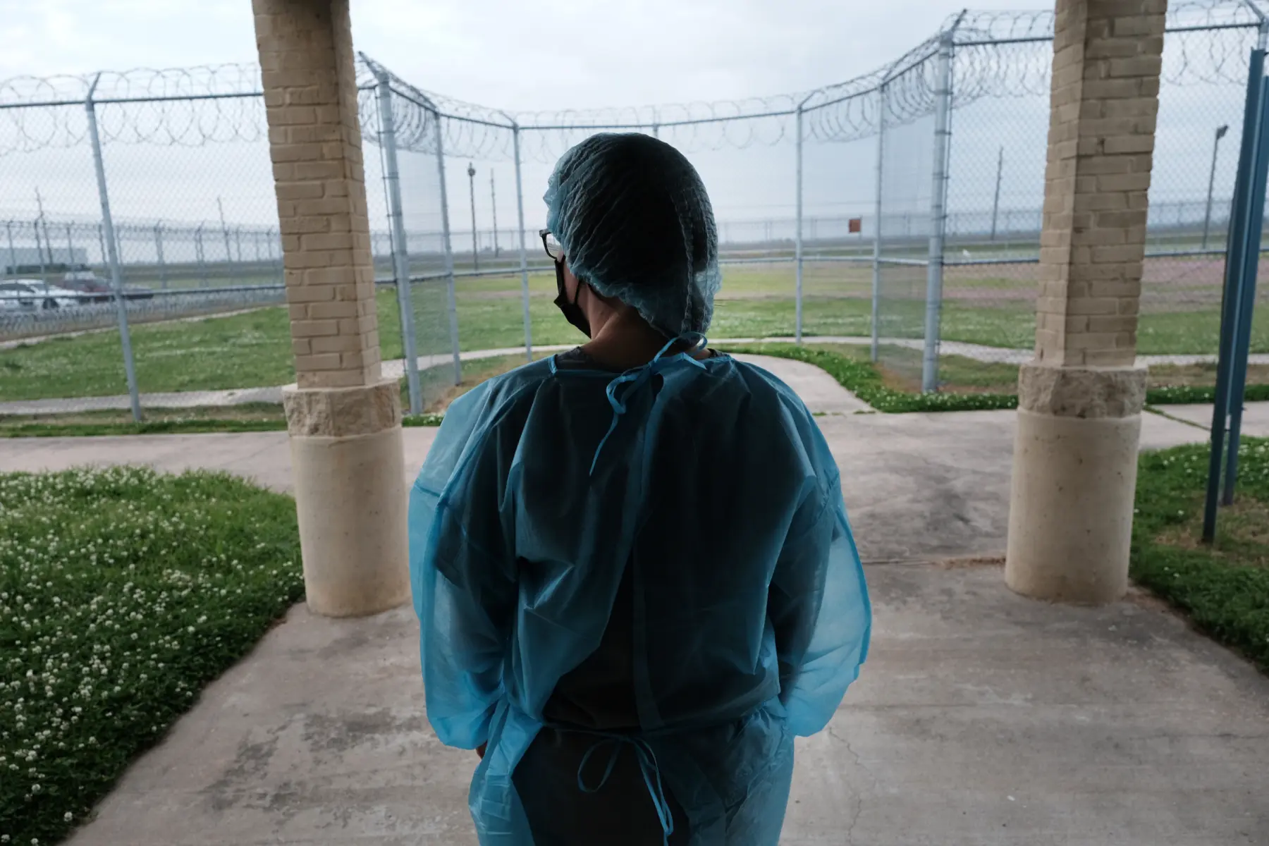A person wearing protective blue medical gear stands at an entrance, gazing out toward a fenced area with grass and barbed wire.