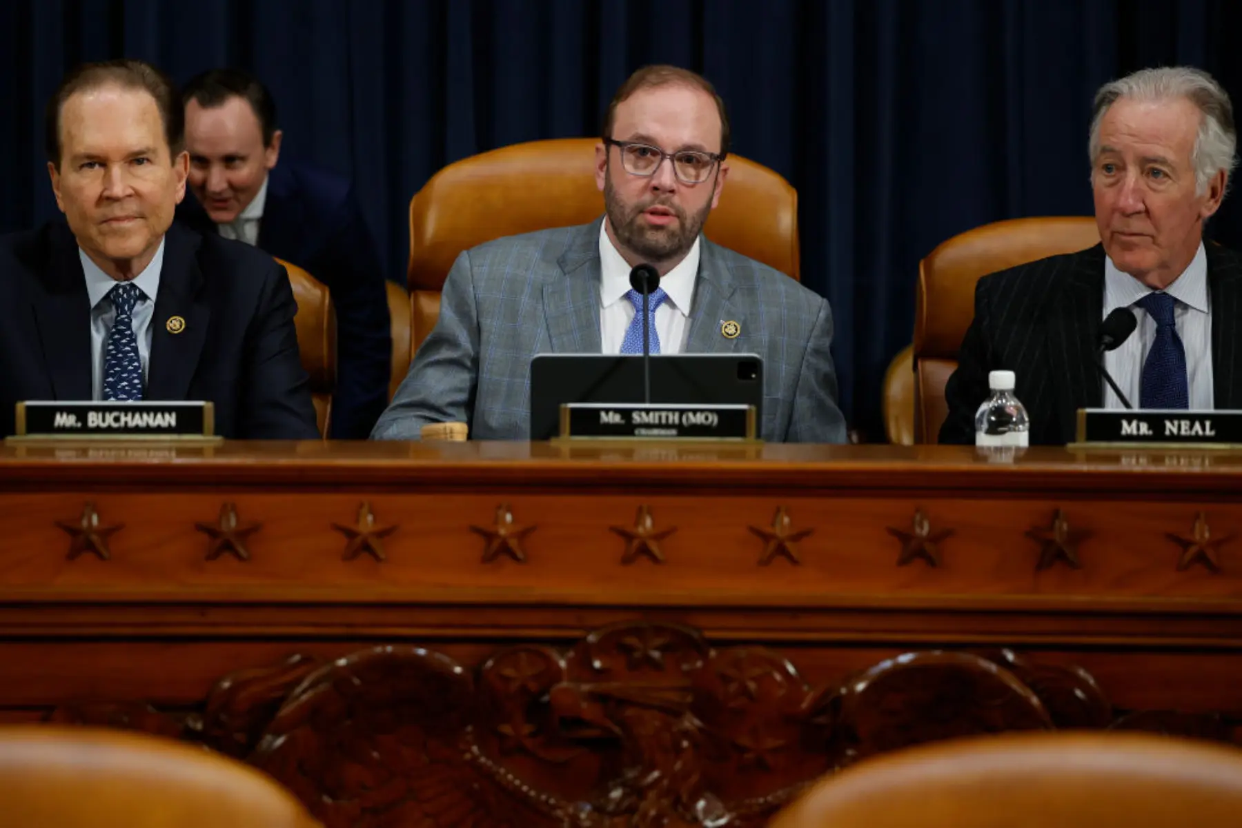 Three men sit at a legislative hearing table, engaged in discussion. A blue curtain serves as a backdrop, with decorative woodwork visible.