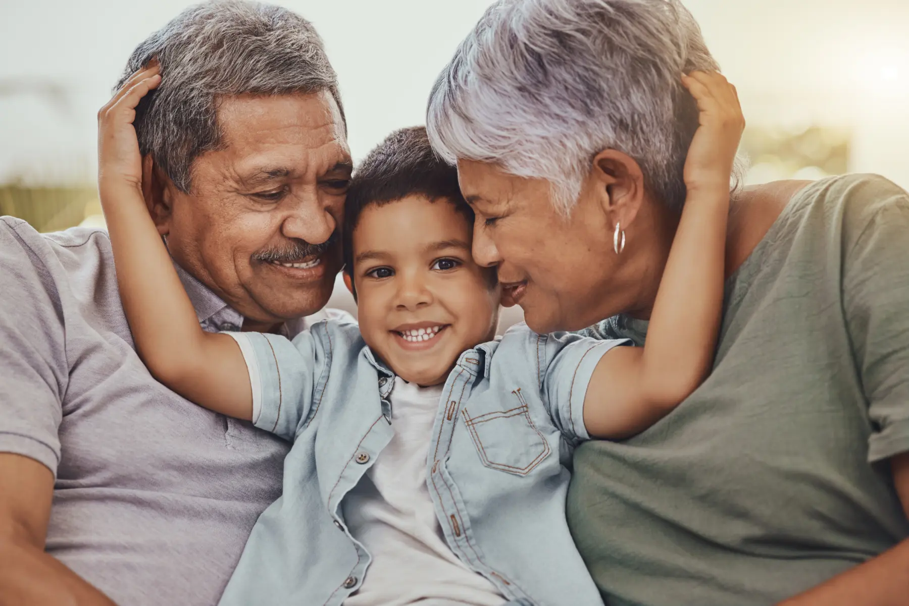 A smiling boy sits between his joyful grandparents, sharing a warm embrace, radiating love and connection in a sunlit setting.