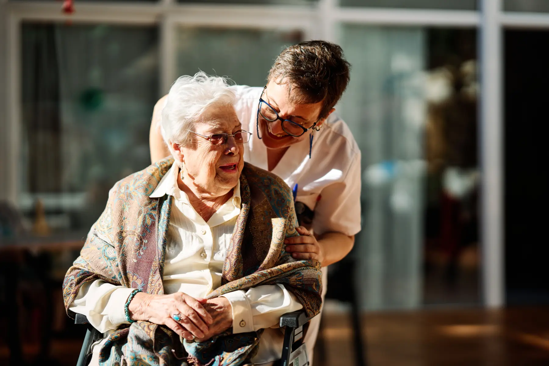 An elderly woman smiles softly while seated, wrapped in a shawl, as a caregiver gently interacts with her in a warm, inviting setting.