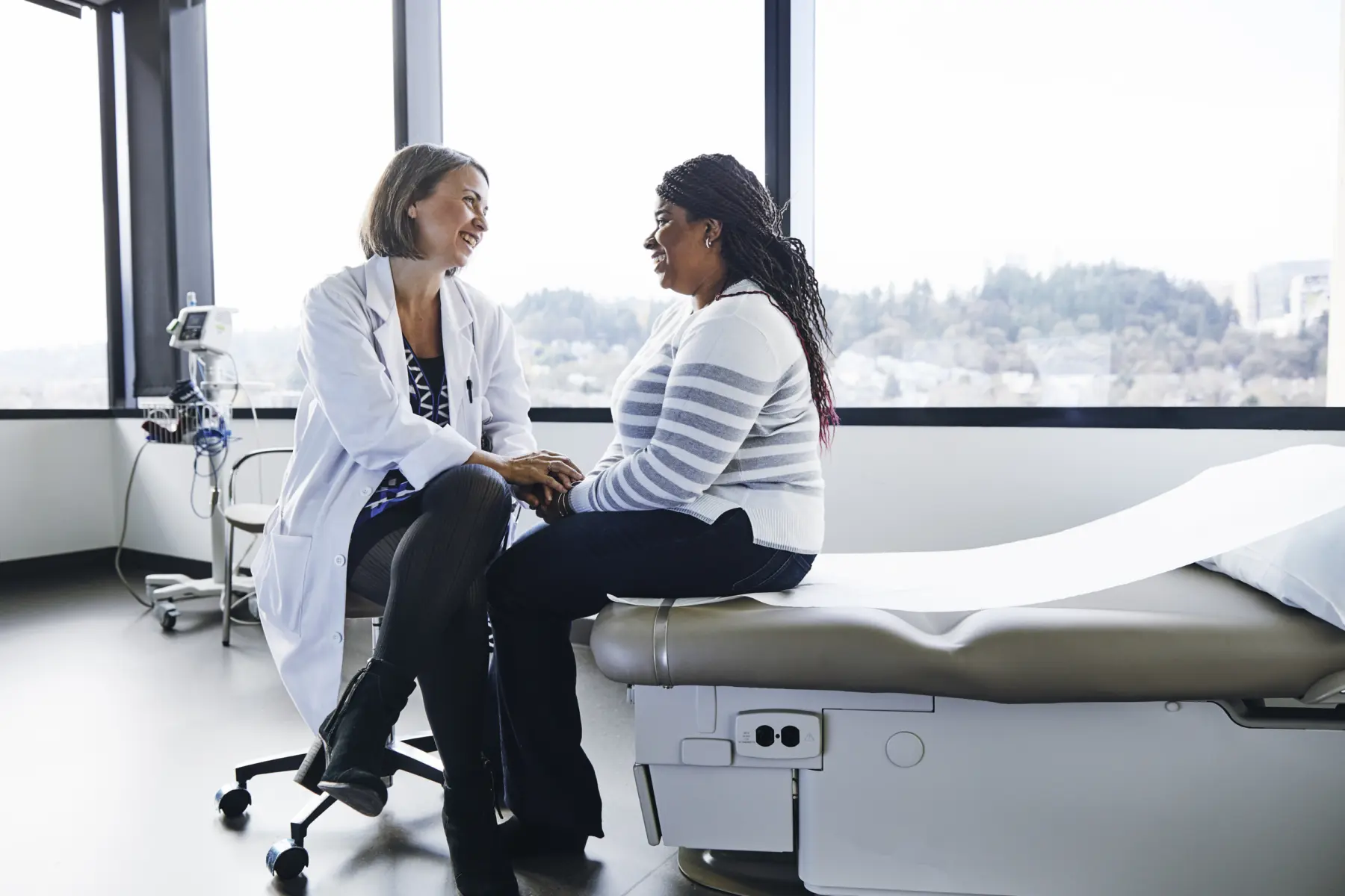 A smiling doctor and patient sit closely, sharing a warm conversation in a bright medical office with large windows.