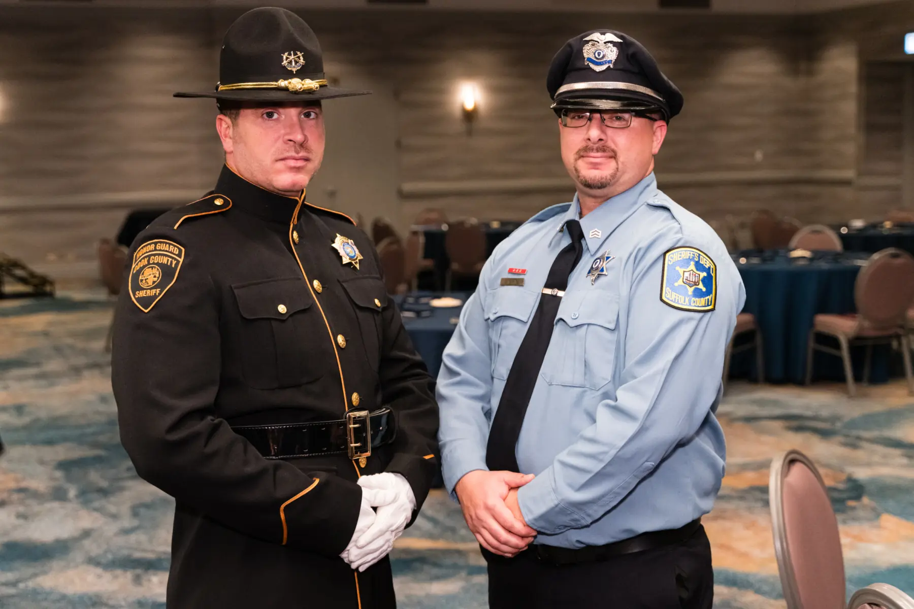 Two law enforcement officers stand together in a banquet hall, one in a formal dark uniform with a hat, the other in a light blue uniform.