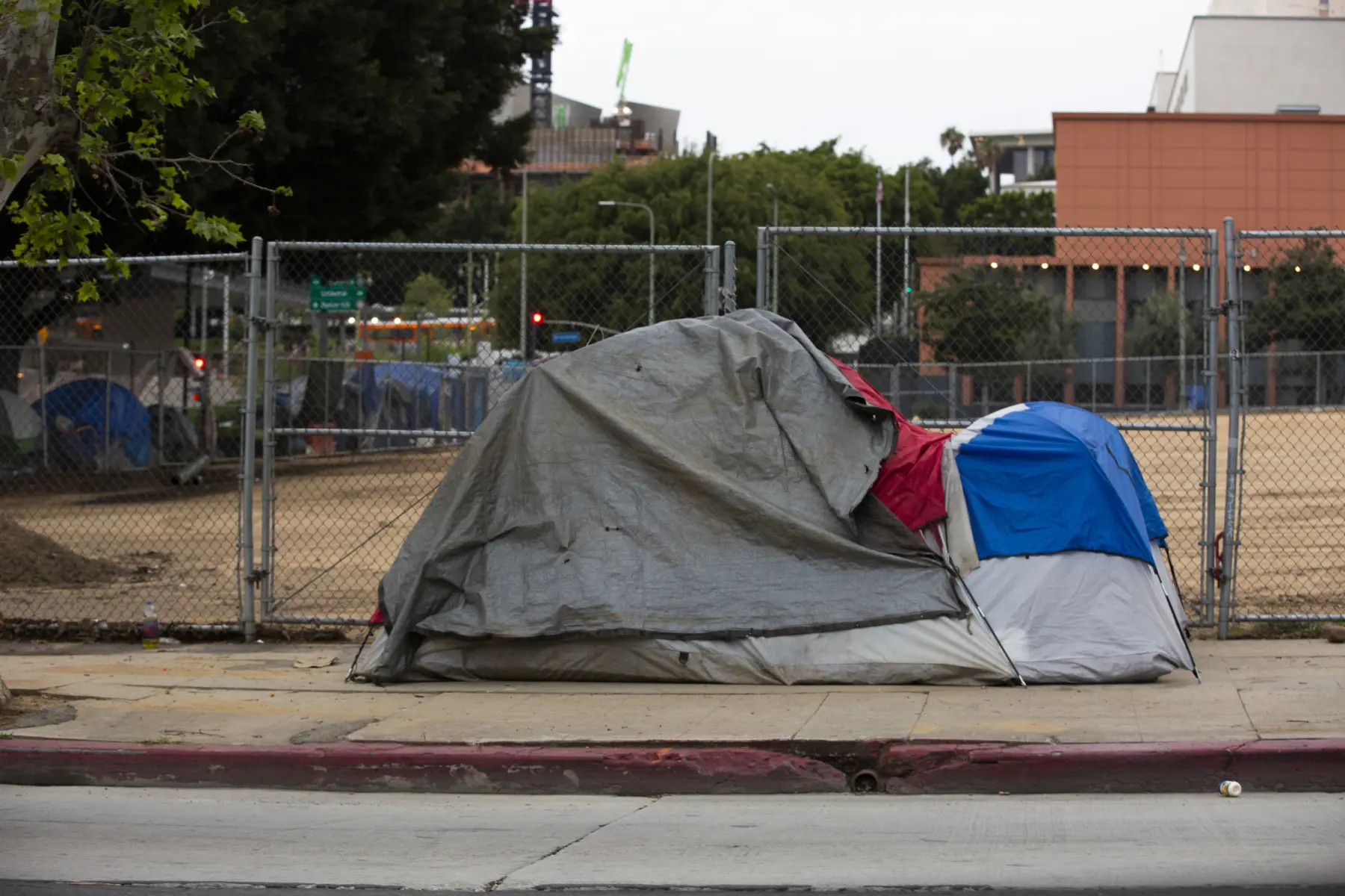 A weathered tent, covered with a gray tarp and touches of red and blue, sits along a sidewalk near a fenced area.