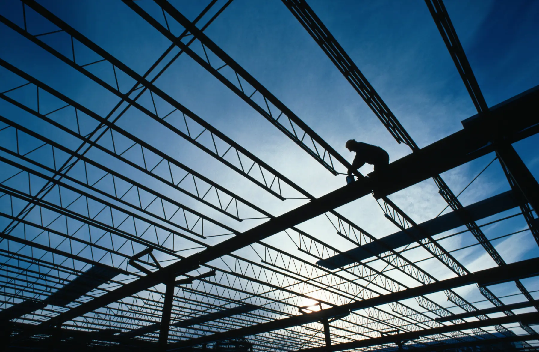 A silhouetted worker balances on a steel beam, gripping a tool, against a blue sky and intricate framework of beams above.