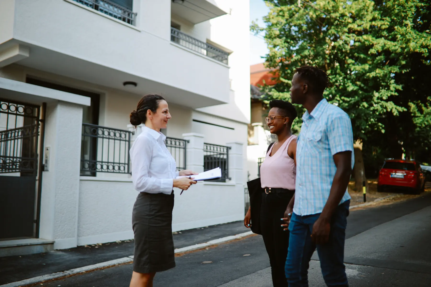 A woman in a white shirt and skirt holds papers while smiling at two people on a street, surrounded by greenery and buildings.