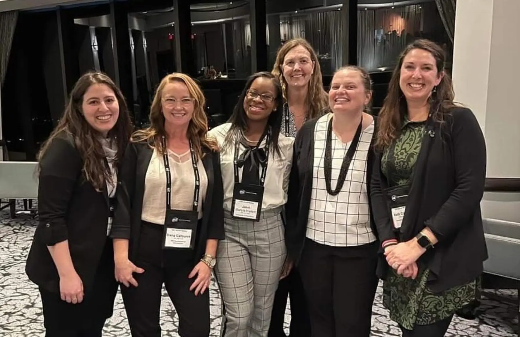 Six women pose together in a conference setting, wearing badges, smiling, and dressed in professional attire against a stylish backdrop.