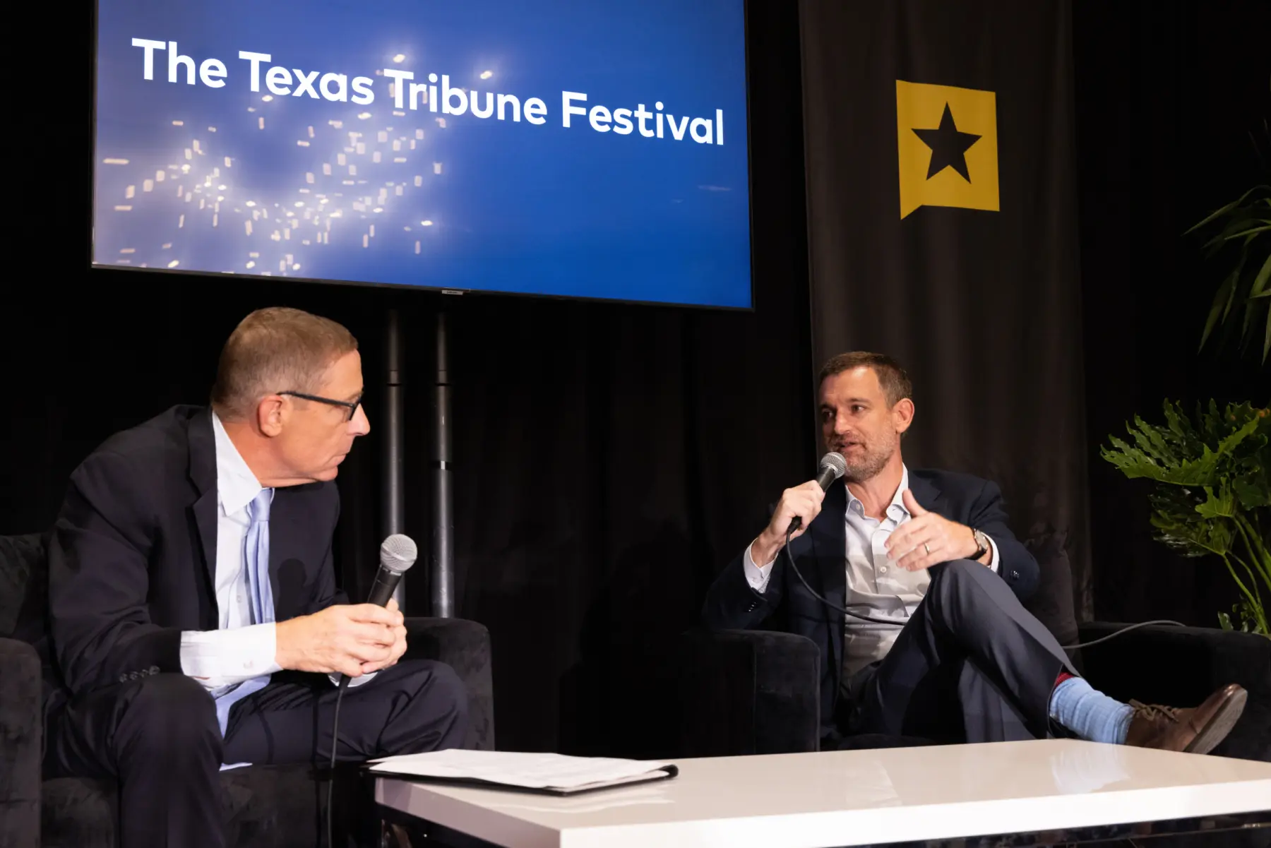 Two men, one speaking into a microphone, engage in conversation on stage at the Texas Tribune Festival, with a spotlighted backdrop.