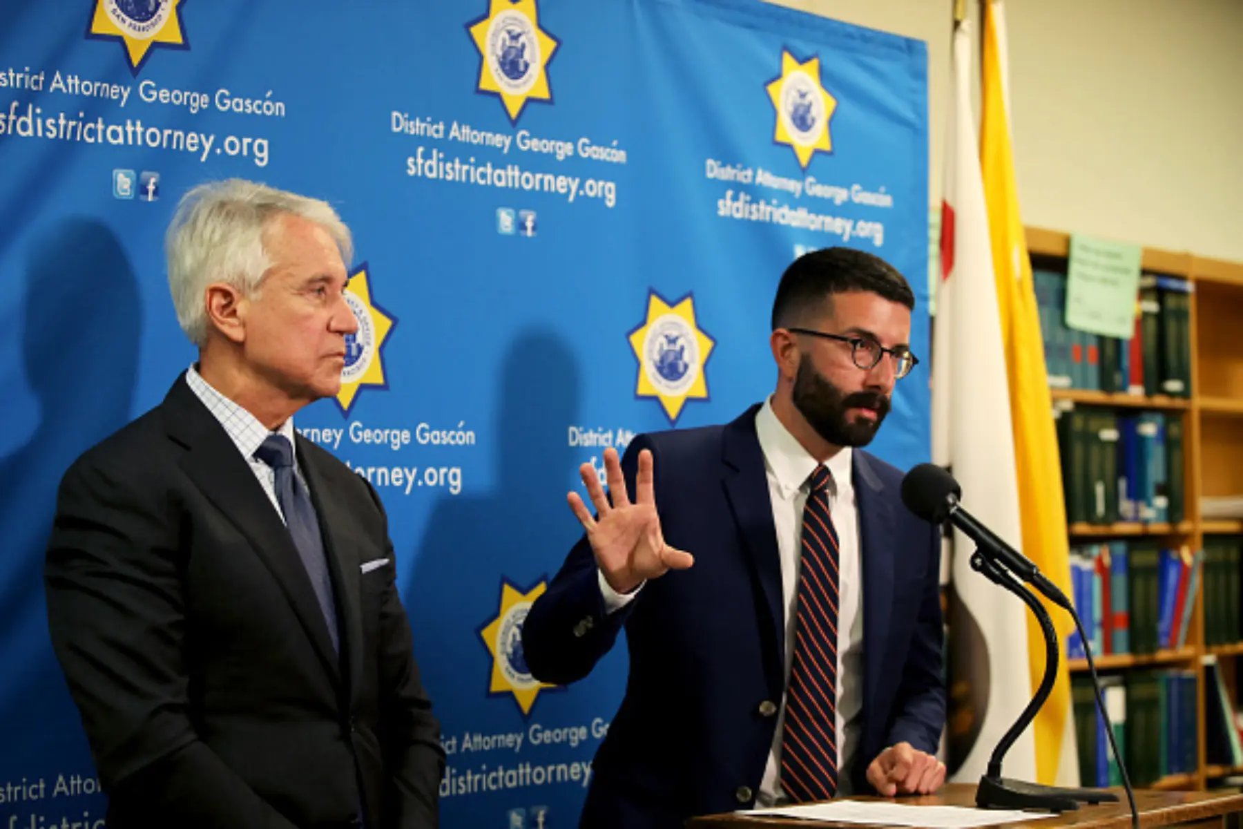 Two men stand at a podium, one gesturing with his hand while the other listens. Behind them is a blue backdrop with law enforcement seals.