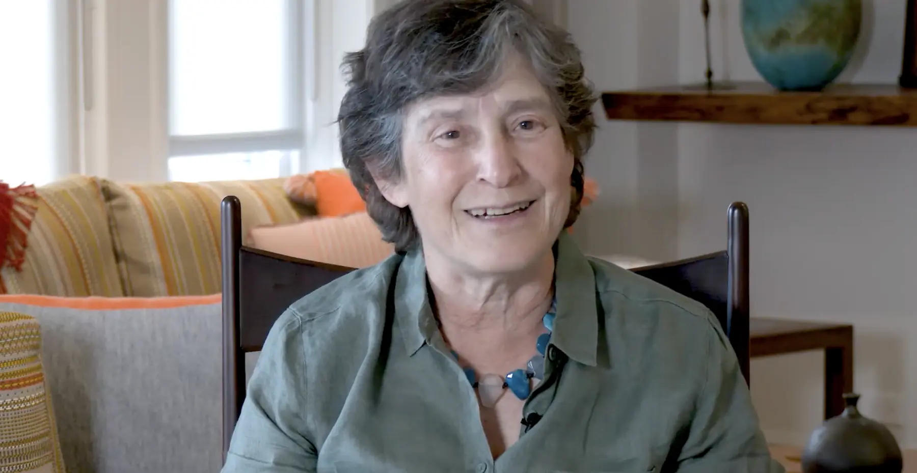 A smiling person with short, wavy hair wears a green shirt and a blue necklace, seated in a cozy room with colorful cushions.