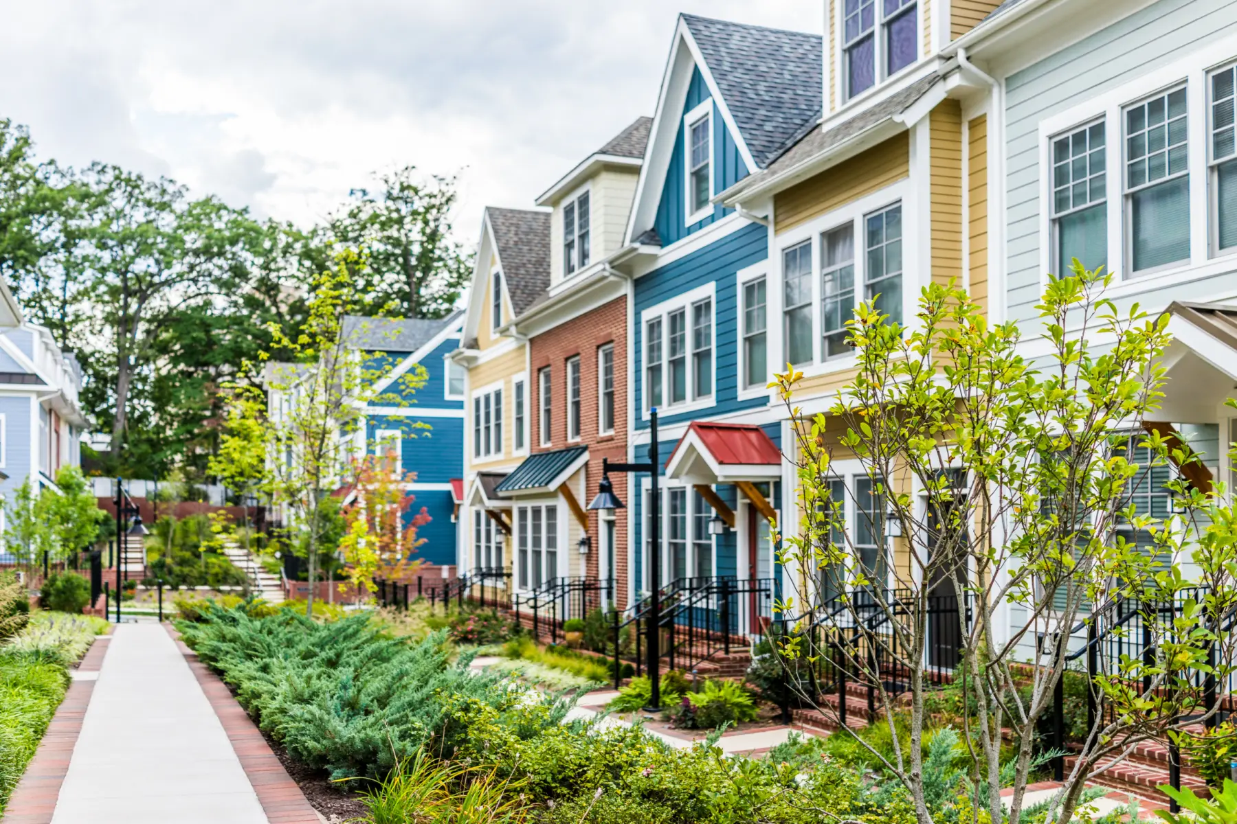 Colorful townhouses line a well-maintained path, surrounded by greenery and blooming plants, under a partly cloudy sky.
