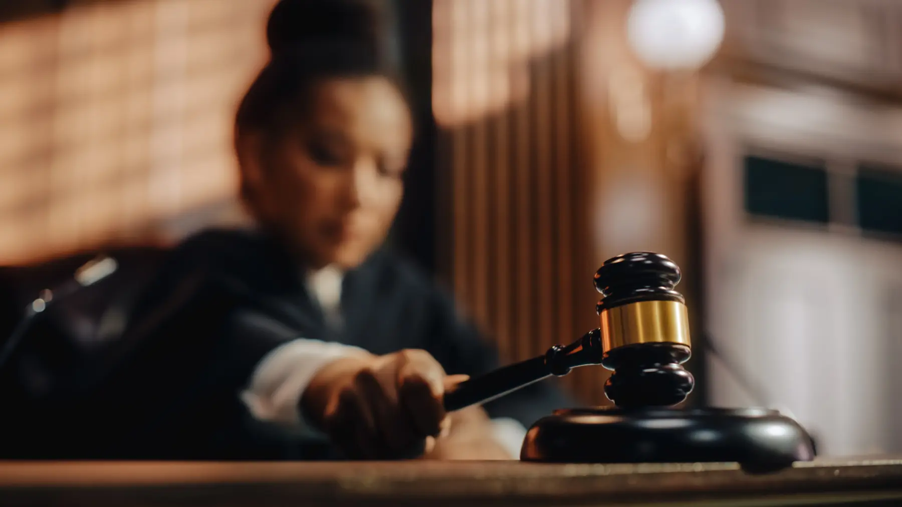 A judge's gavel rests on a table in a courtroom, while a blurred figure in judicial robes prepares to bring it down decisively.