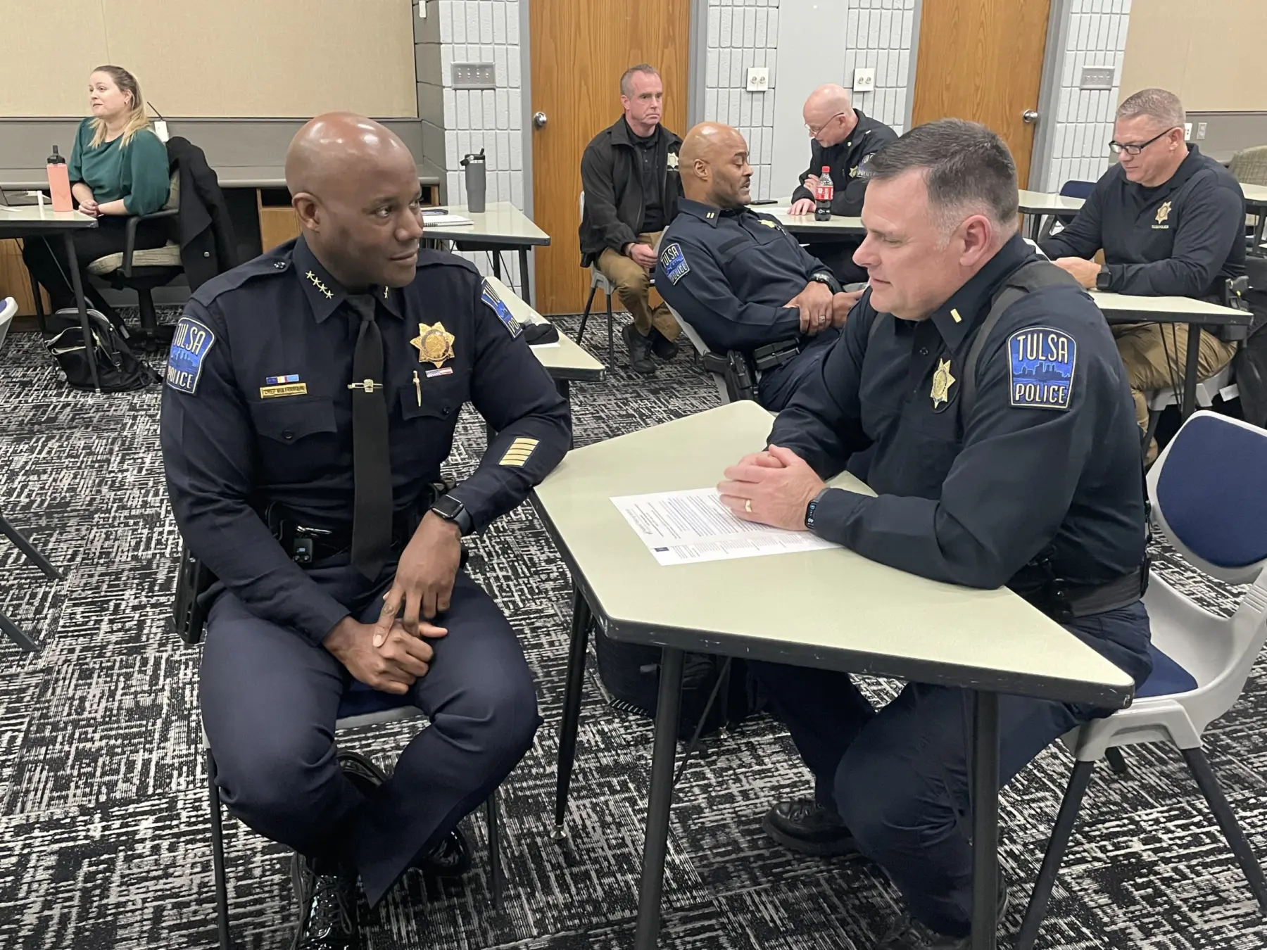Two police officers in uniform sit at a table engaged in a conversation, while colleagues are seated in the background, focused on tasks.