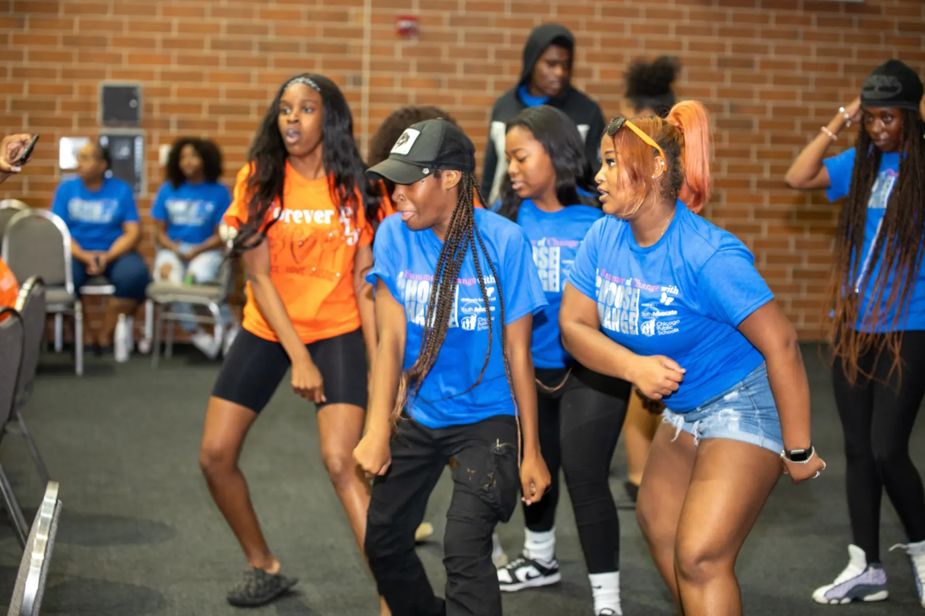 A group of young women in colorful t-shirts dance energetically in a lively indoor setting.