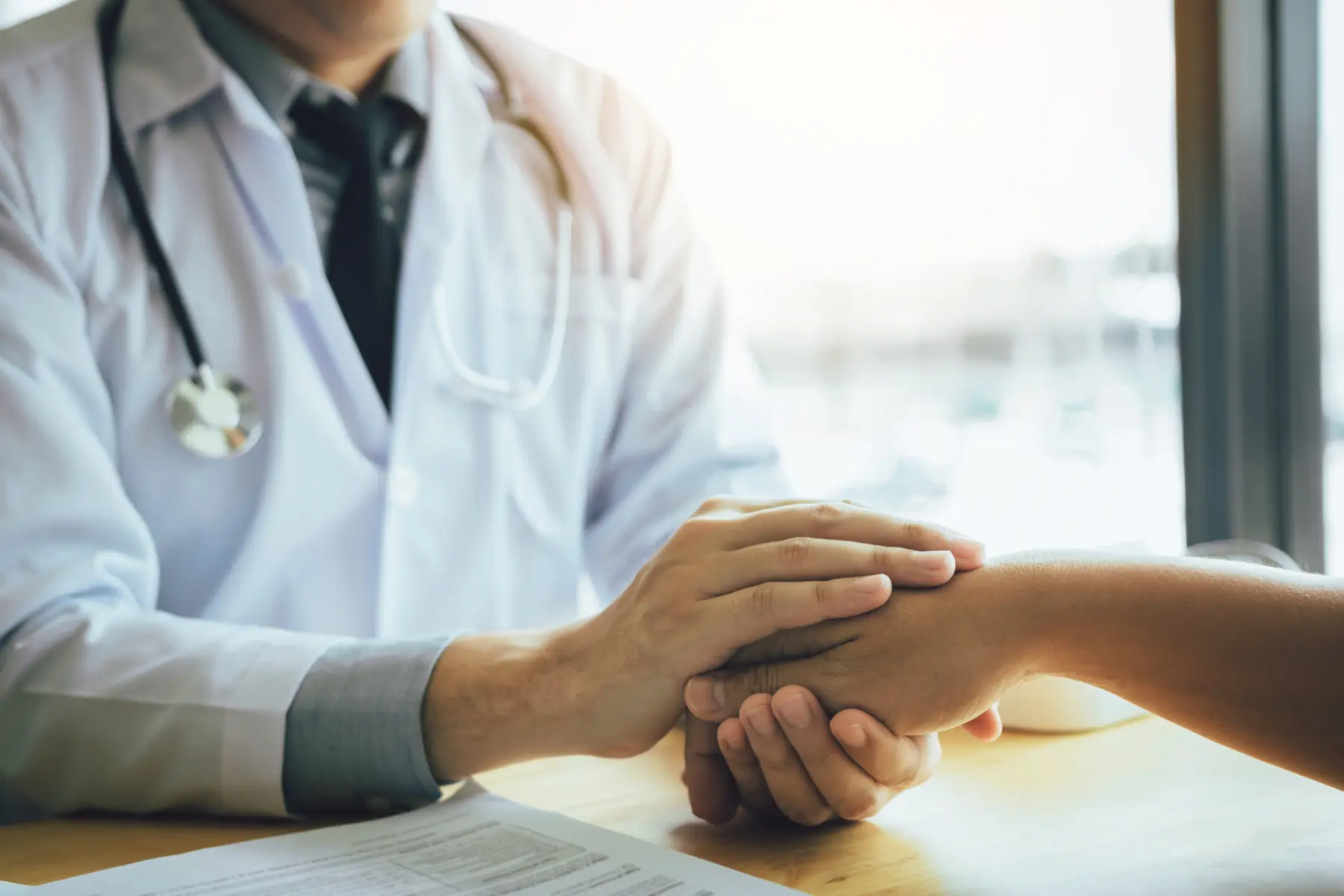 A doctor and patient are holding hands across a table, conveying comfort and support during a medical consultation.