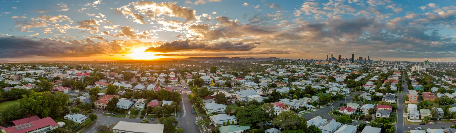 A vibrant sunset illuminates a suburban neighborhood, with colorful houses and trees, framed by distant city skyscrapers and rolling hills.