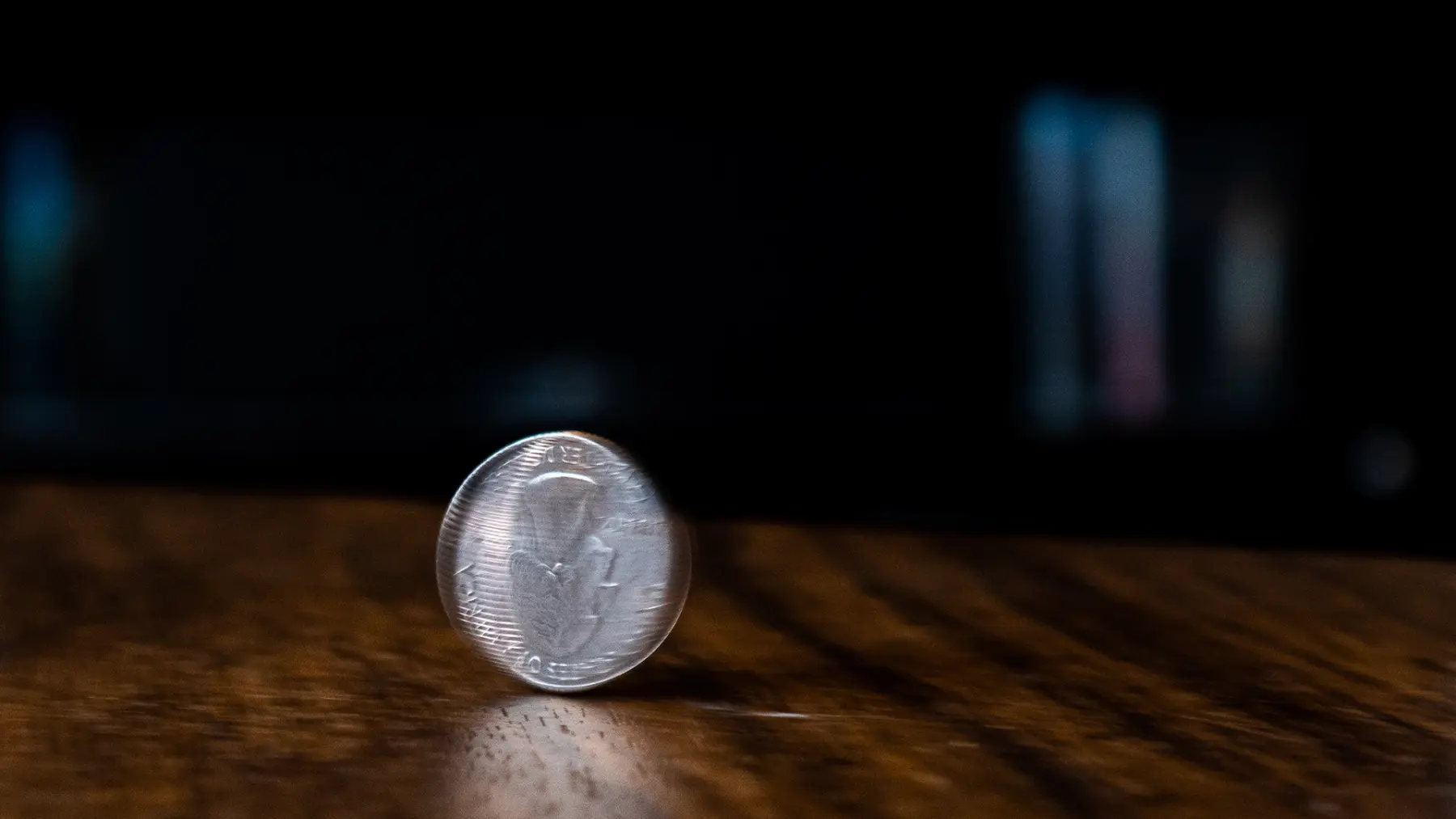 A shiny coin stands upright on a wooden surface, spinning, with a blurred dark background enhancing its gleaming surface.