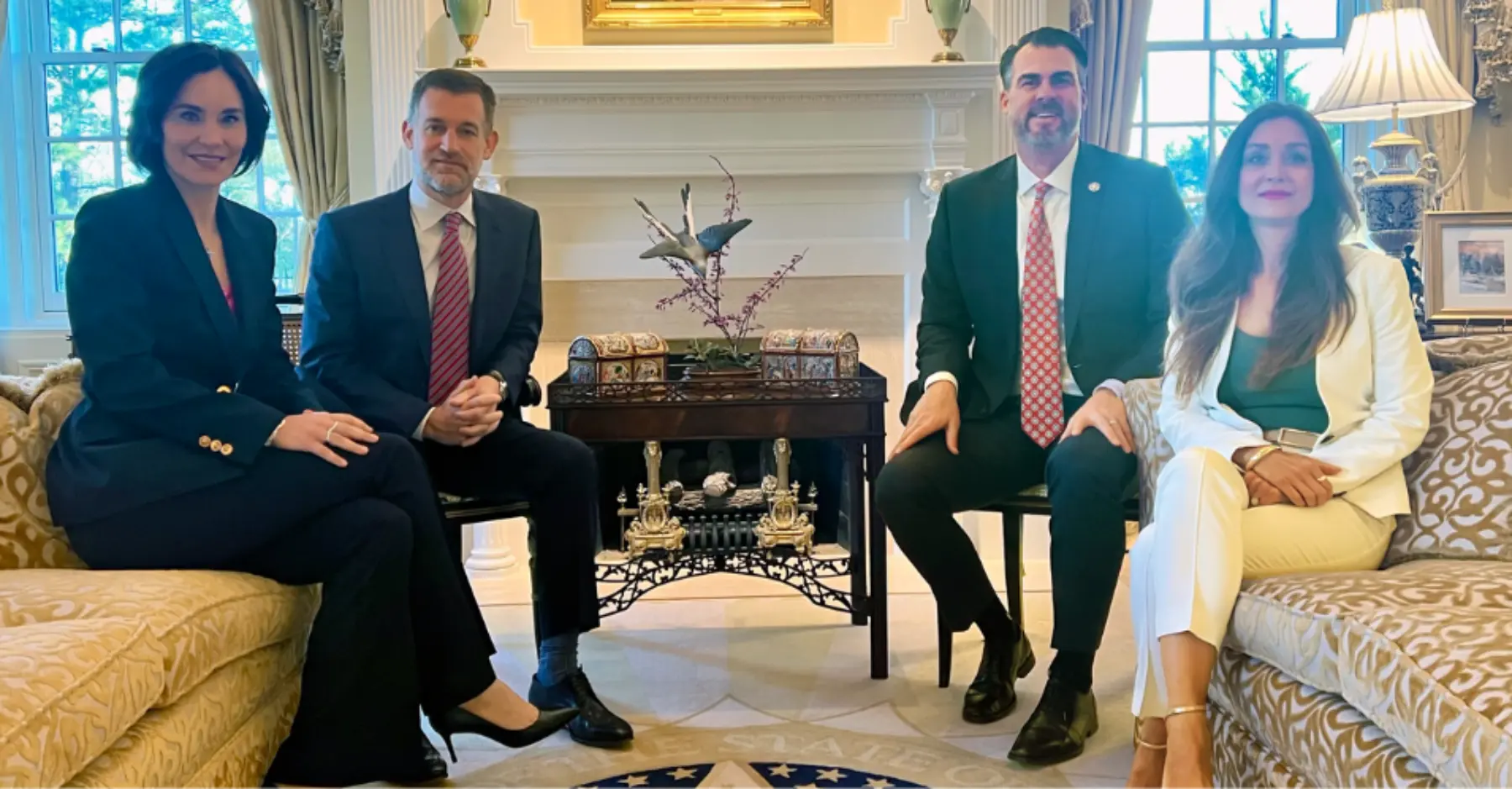 Four people dressed in business attire sit together in an elegant room, with a decorative table and a fireplace in the background.