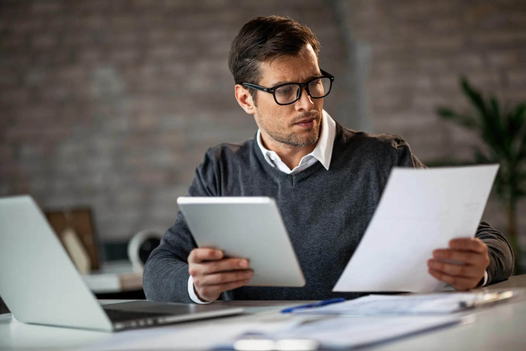 A man in glasses examines a document while holding a tablet, seated at a desk with a laptop and papers in an office.